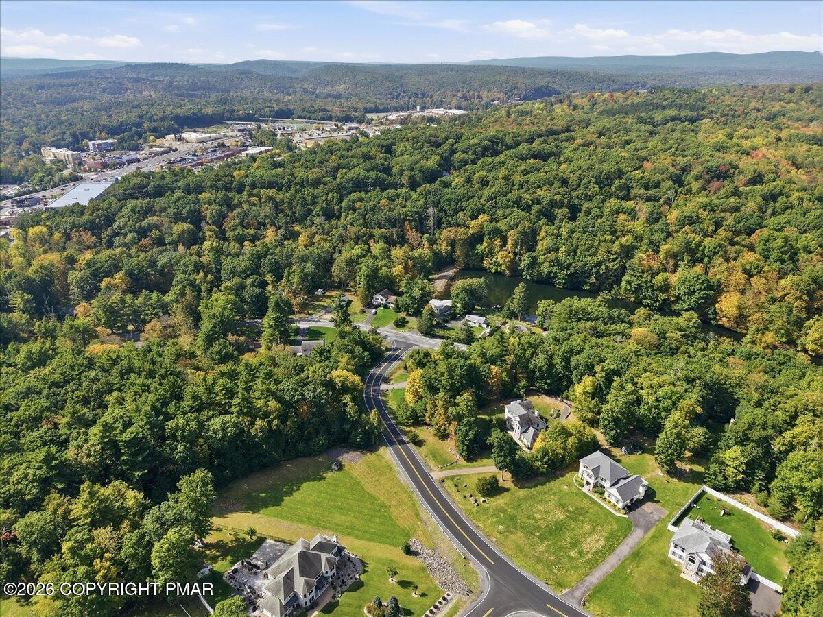 Aerial view of homes among autumn woods, with a winding road, pond, and distant hills.
