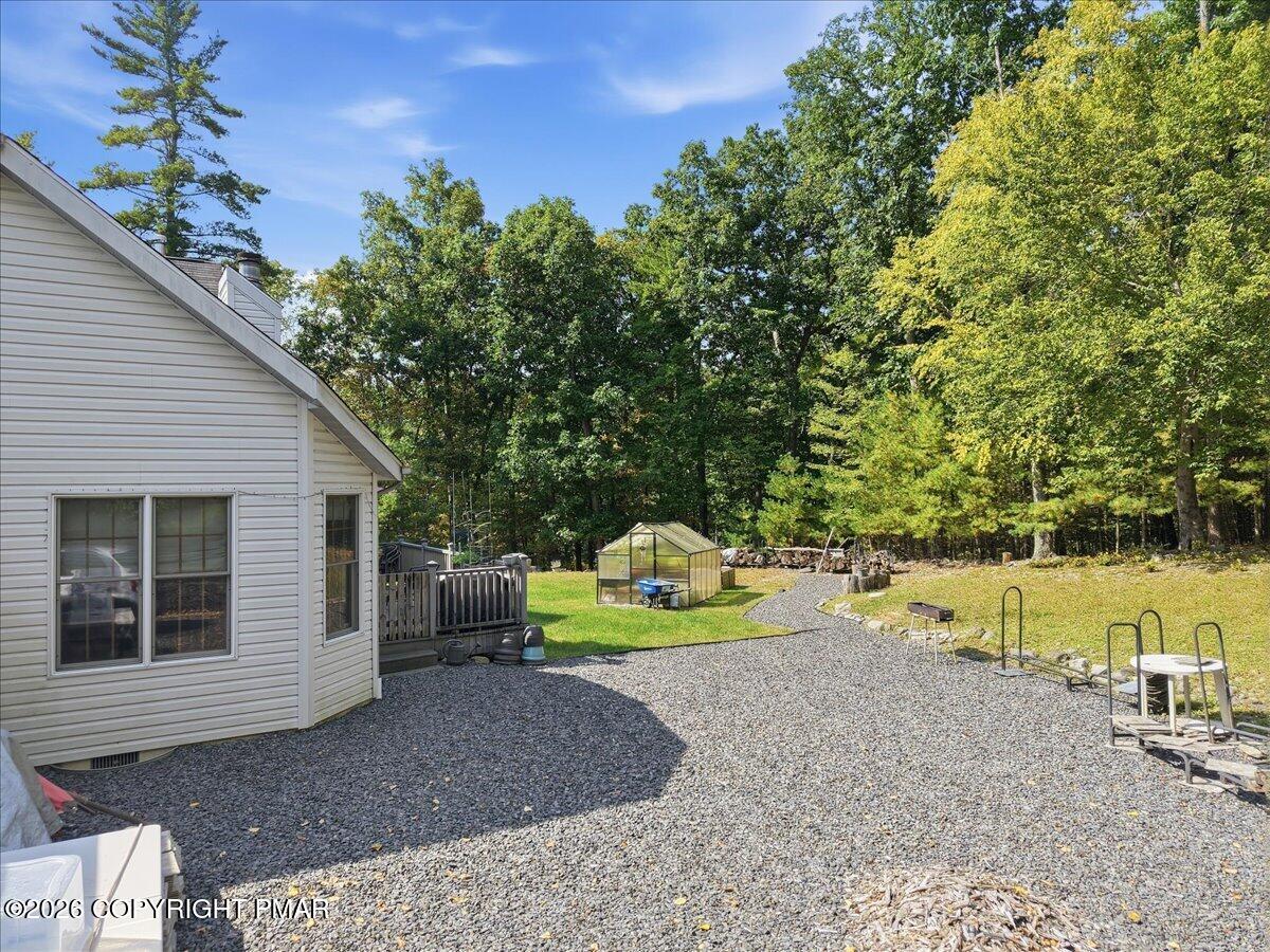 Wooded backyard with a gravel patio, small greenhouse, and deck beside a light-sided house.