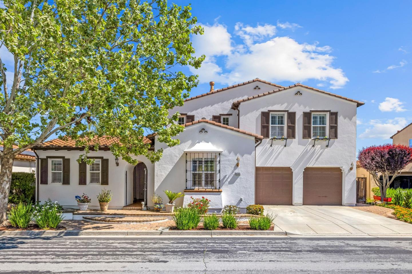 Two-story white stucco home with terracotta roofs, brown shutters, and a two-car garage beneath a leafy tree.