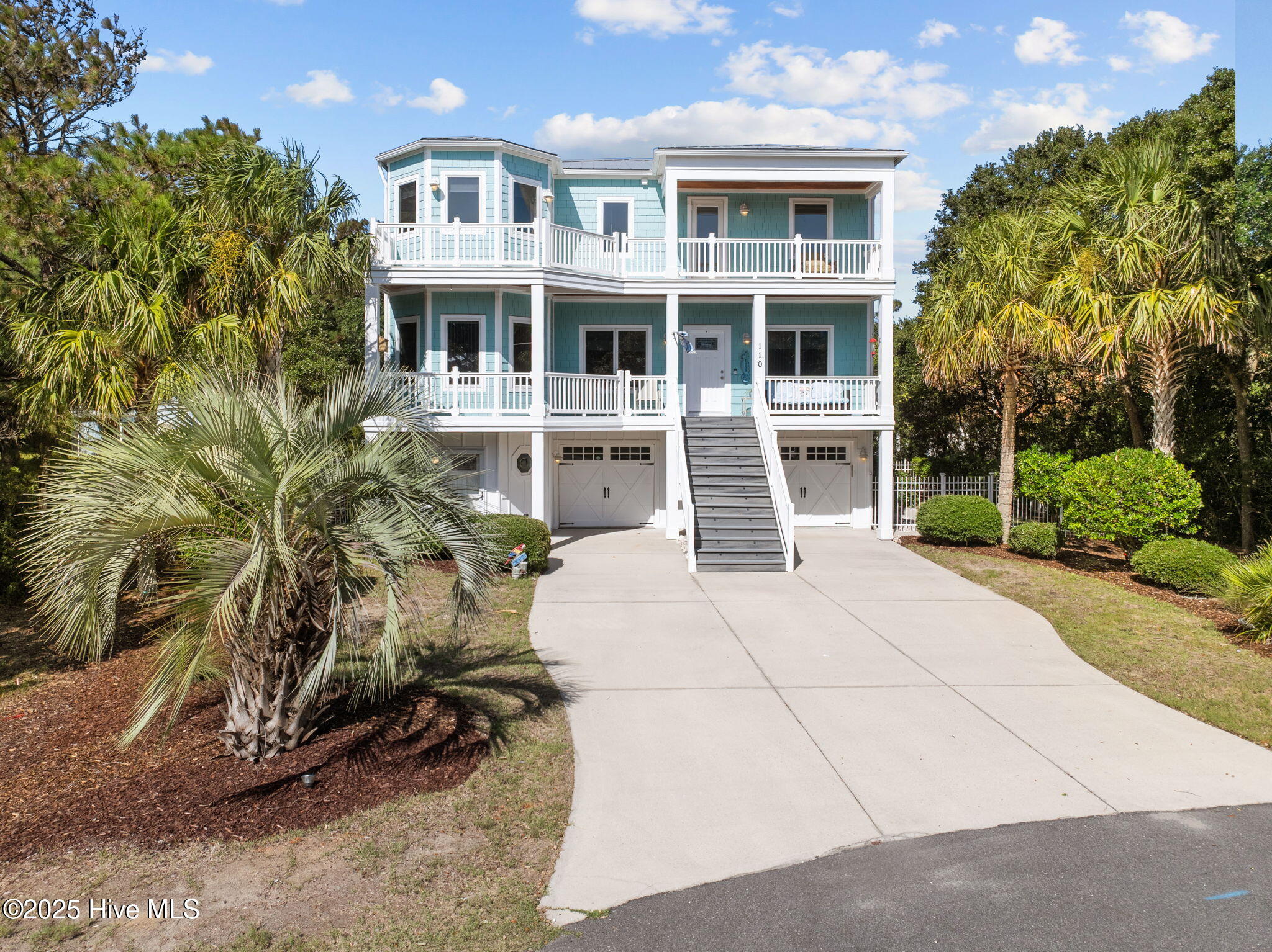 Turquoise coastal home with white balconies, a central staircase, and palm trees along the driveway.