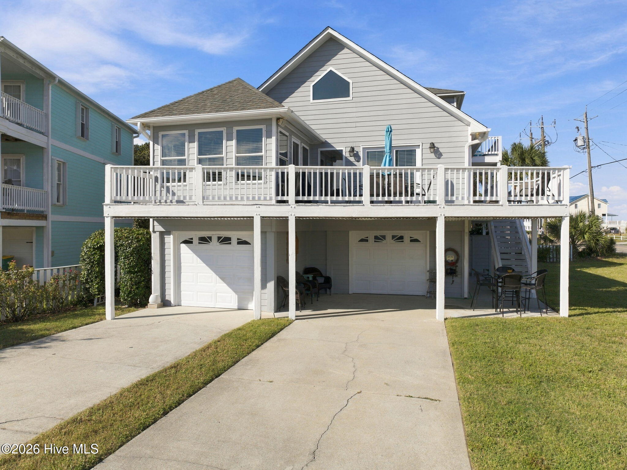 Raised gray coastal house with a broad white-railed deck and two garage doors.