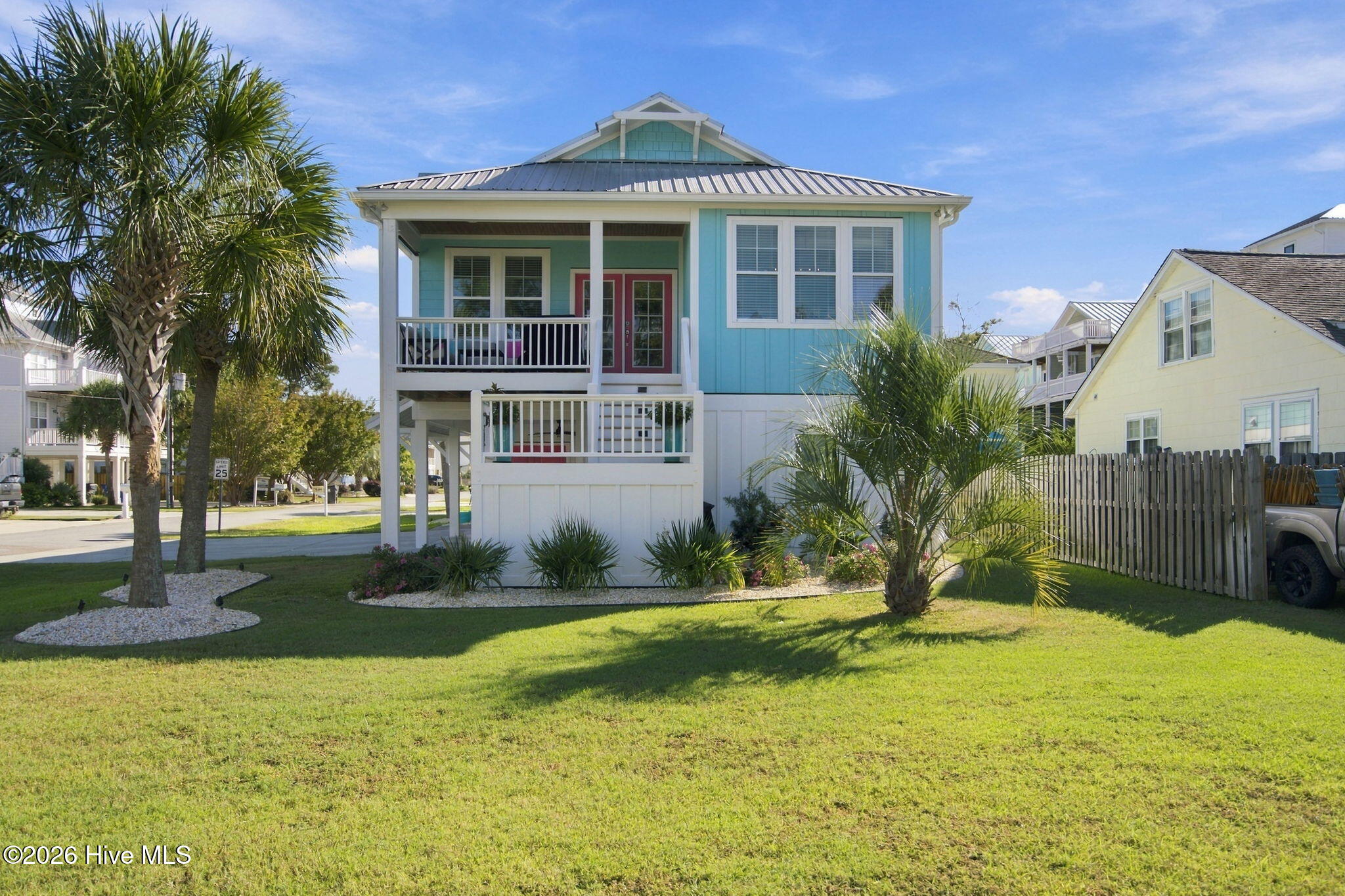 Raised aqua coastal house with white porches, metal roof, and palm trees in the lawn.