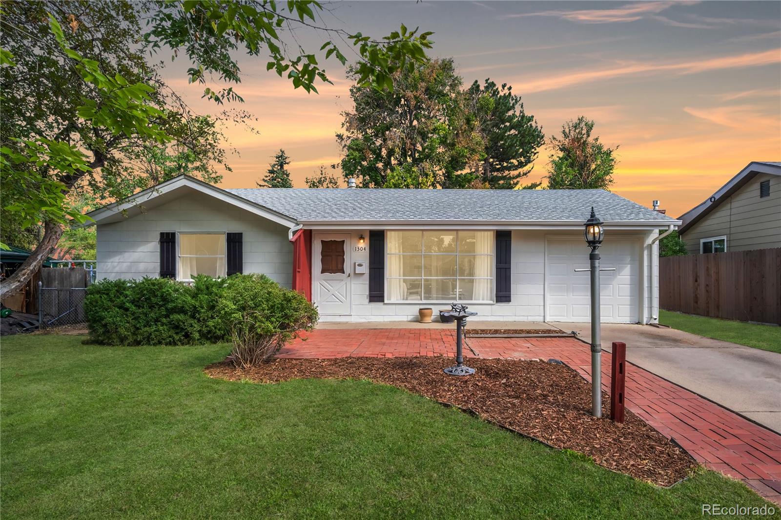 Single-story home with white siding, red-brick walkway, attached garage, and landscaped front yard at sunset.