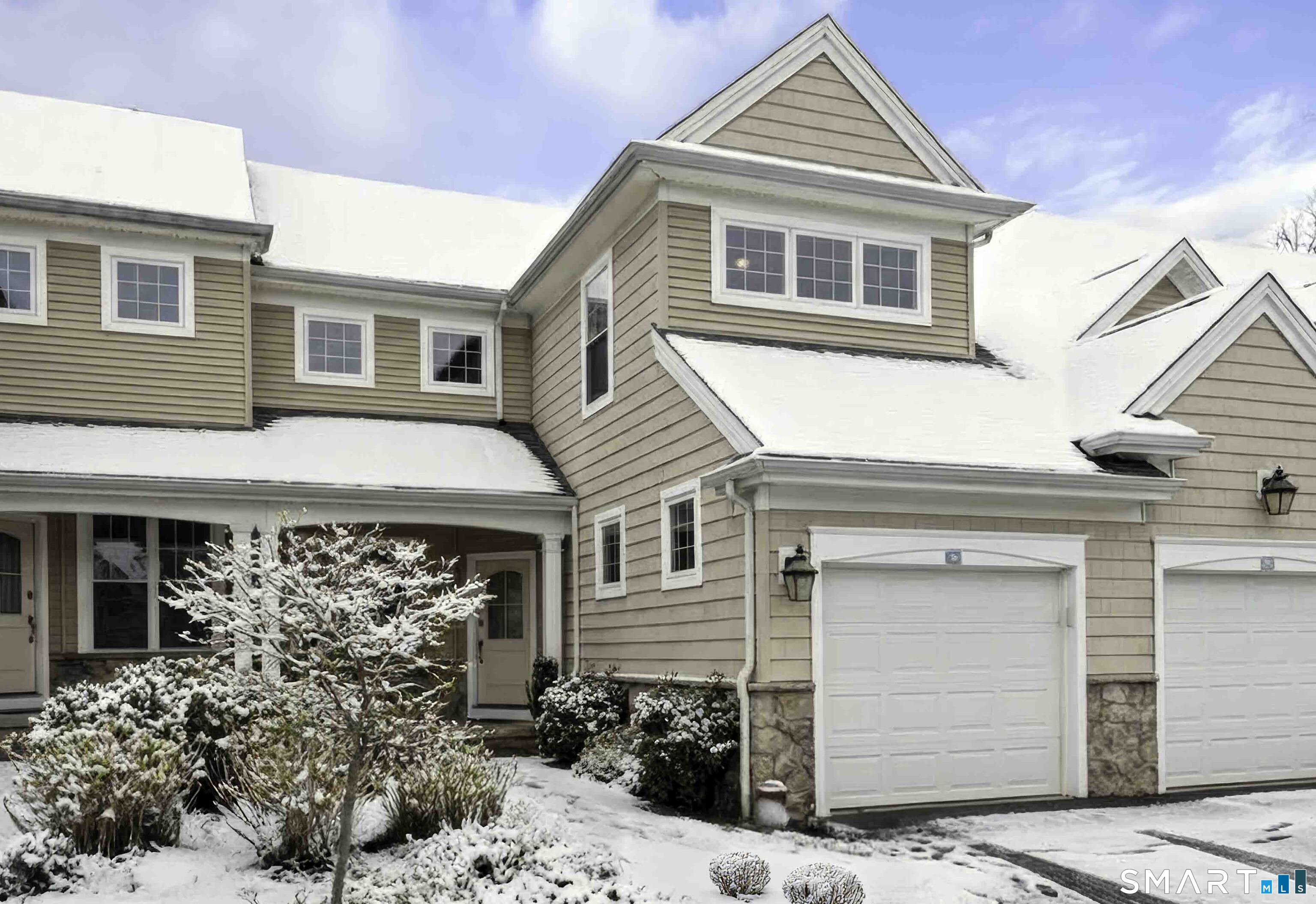 Beige multi-story home with two garage doors and snow-covered roofs and landscaping.