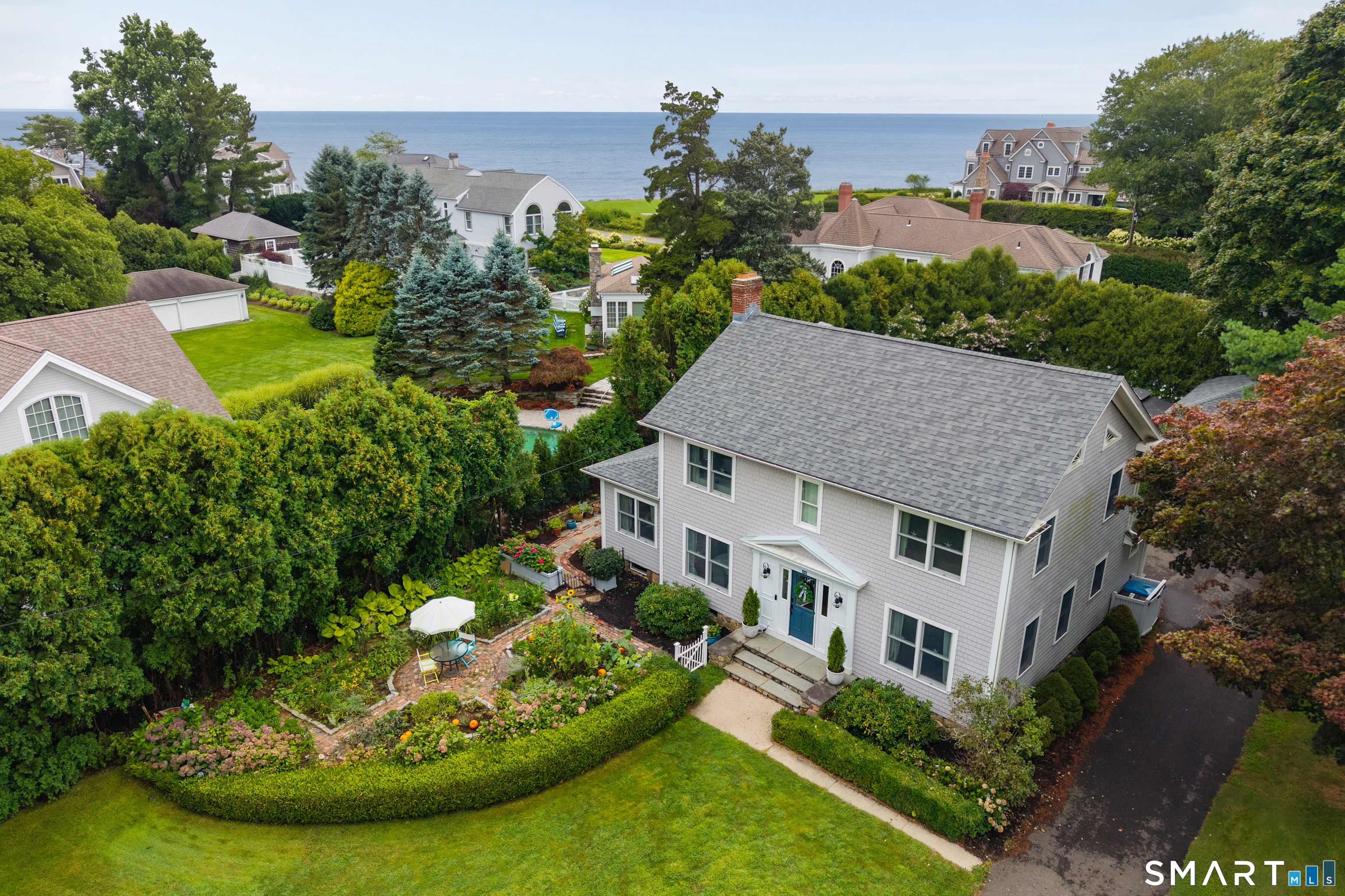 Aerial view of a gray two-story home with a lush garden and shoreline water beyond.