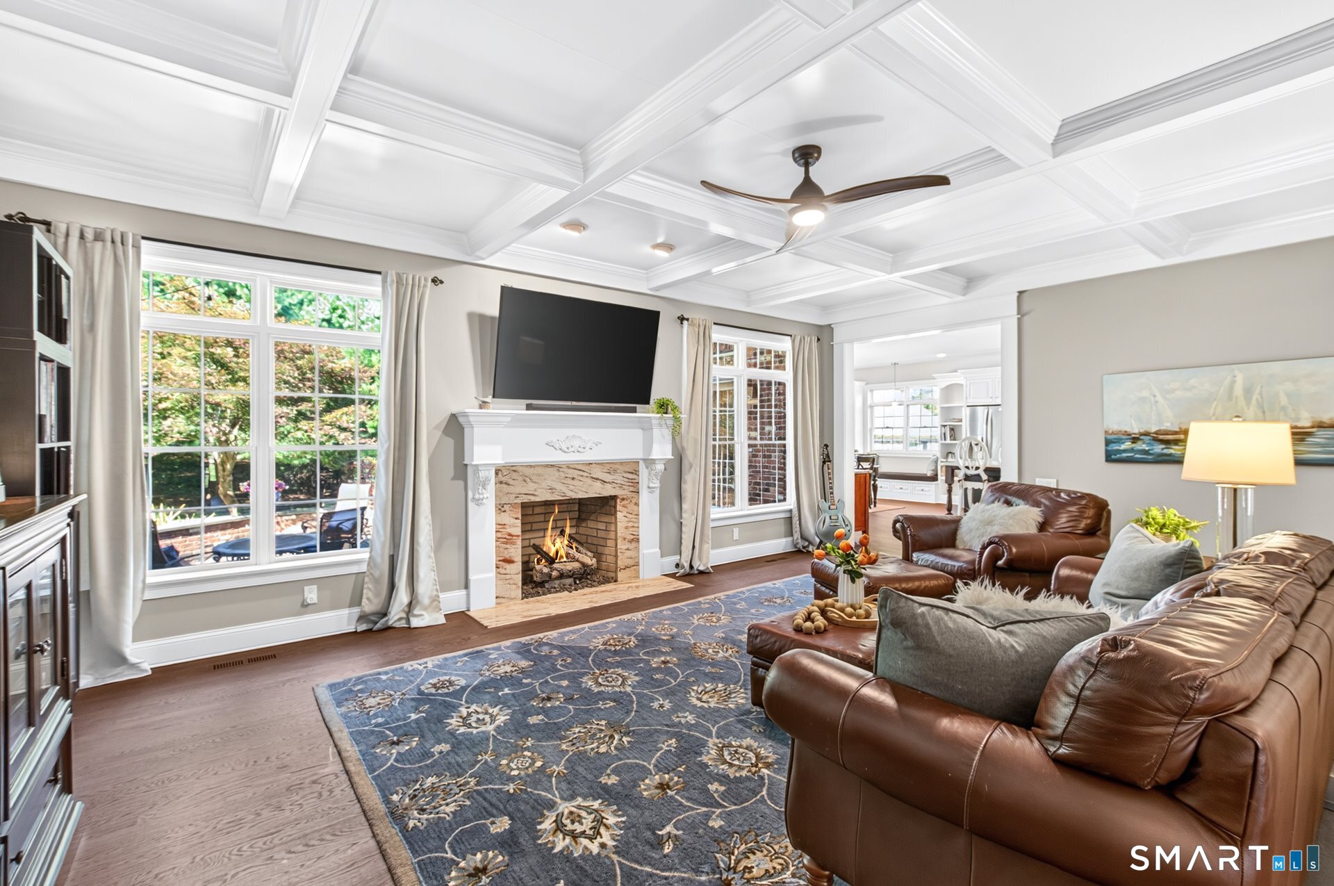 Living room with a coffered ceiling, lit fireplace, large windows, and brown leather seating.