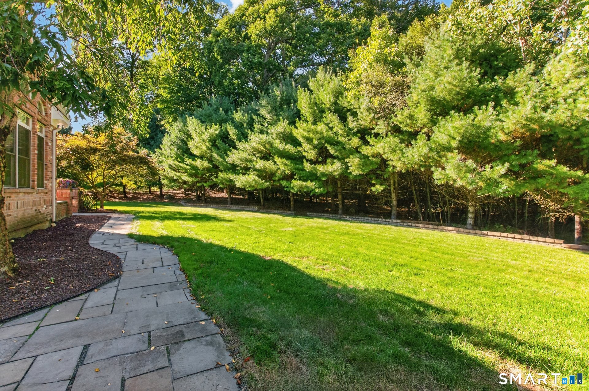 Backyard with a broad lawn, curved stone walkway, brick exterior, and mature evergreen trees.