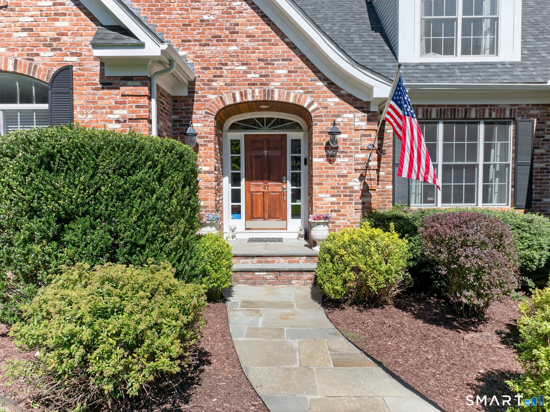 Brick home entrance with an arched doorway, American flag, landscaped shrubs, and stone walkway.
