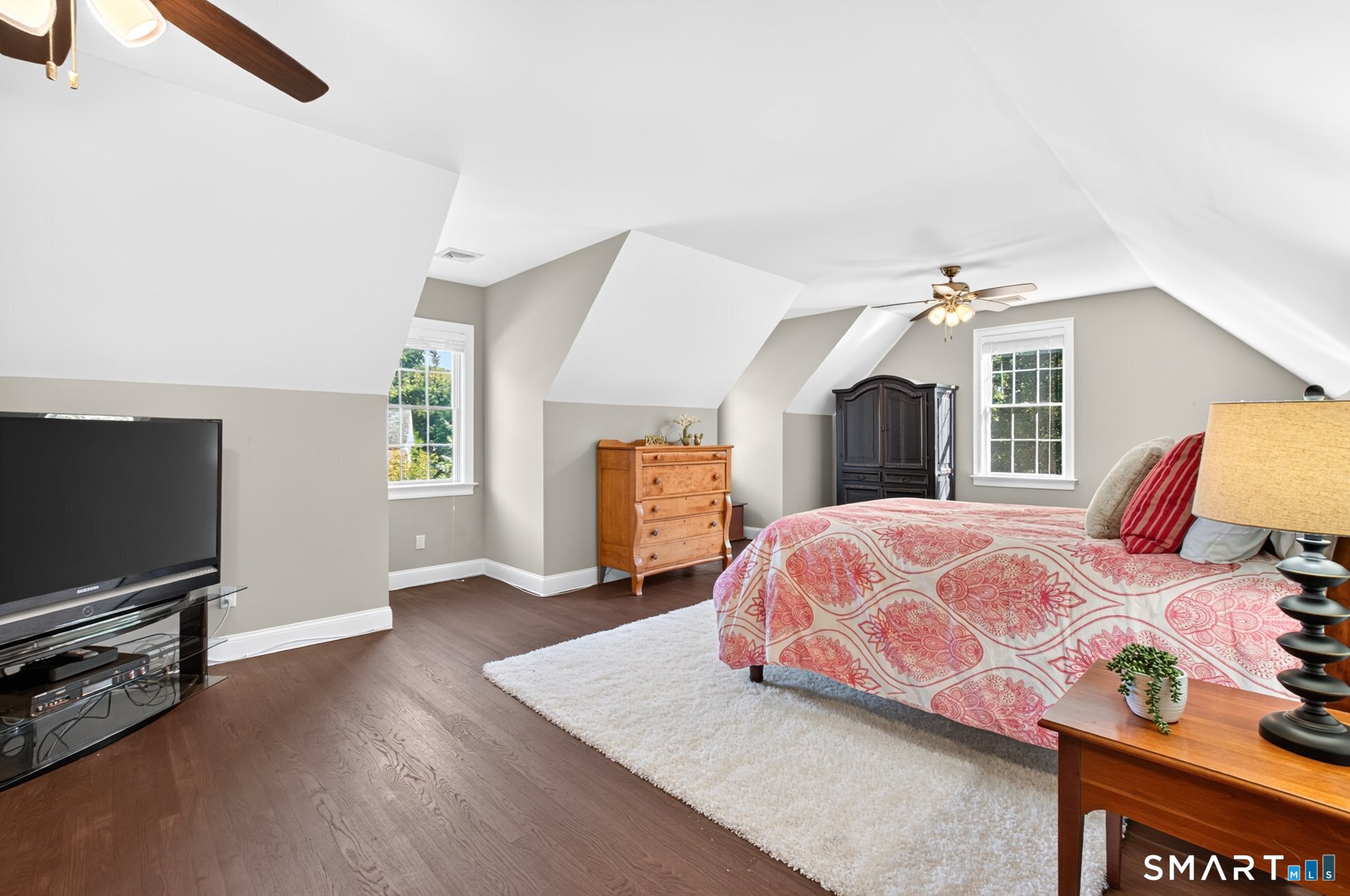 Furnished bedroom with sloped ceilings, a pink patterned bedspread, hardwood floors, and two ceiling fans.