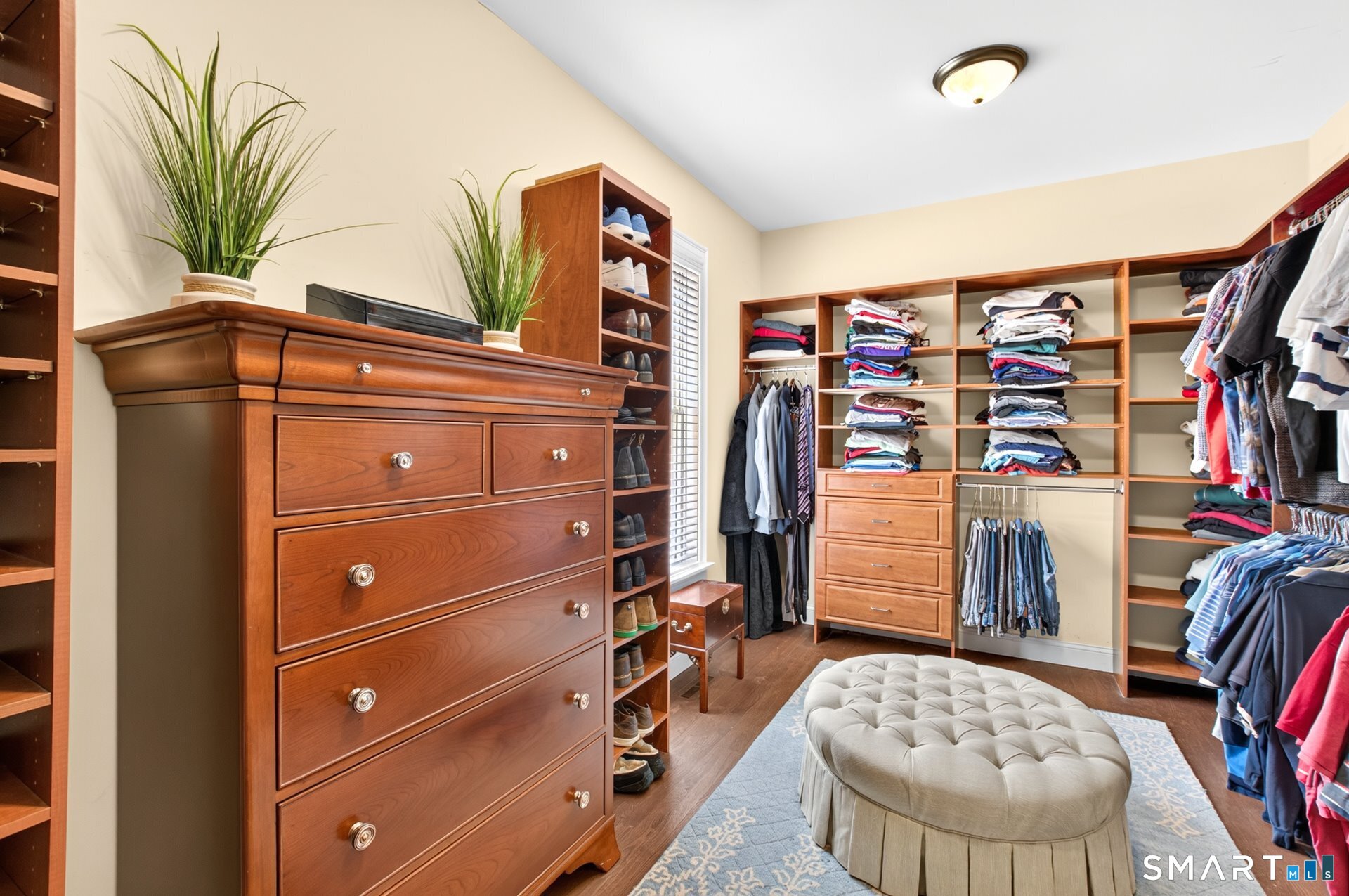 Walk-in closet with extensive wooden built-ins, neatly arranged clothes, and a round tufted ottoman.