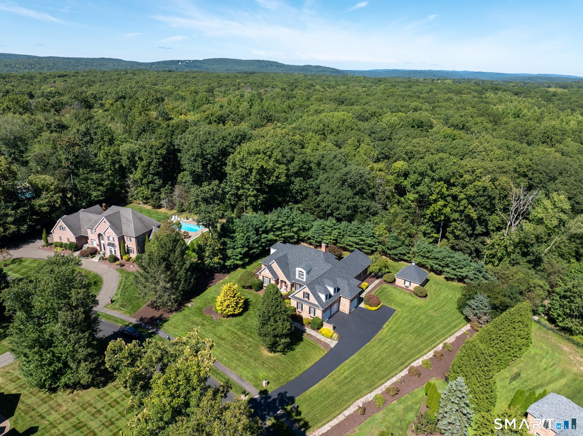 Aerial view of a brick home with a pool, landscaped lawn, and surrounding woodland.