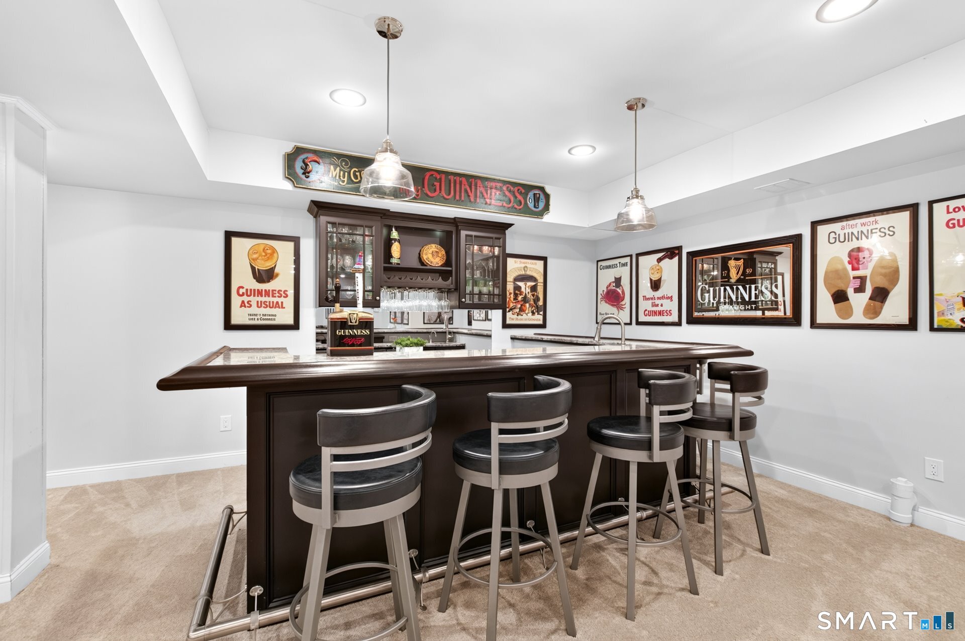 Home bar with a dark wood counter, four stools, glass cabinets, and Guinness-themed wall art.