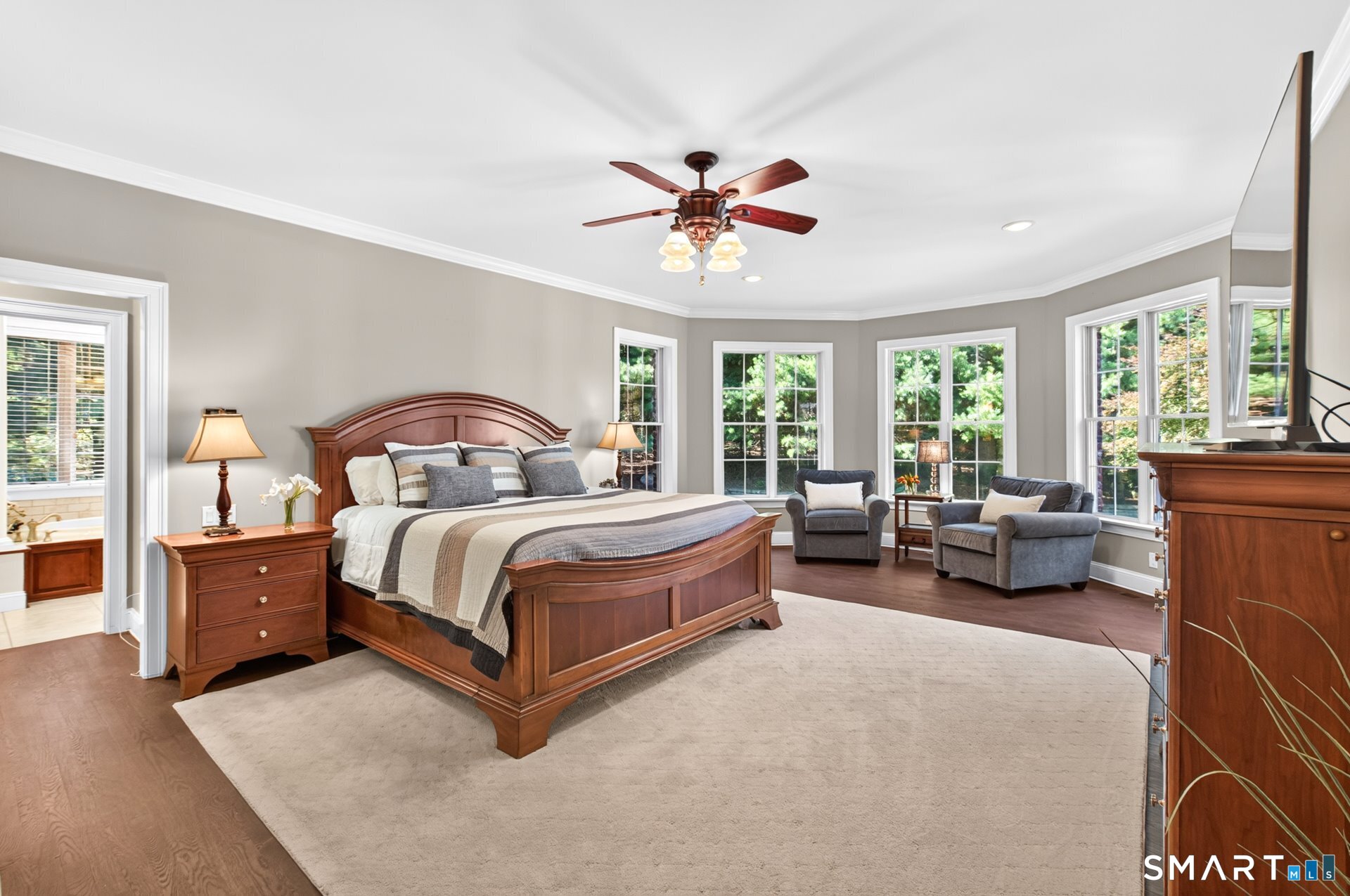 Furnished bedroom with a large wooden bed, sitting area, and tall windows overlooking greenery.