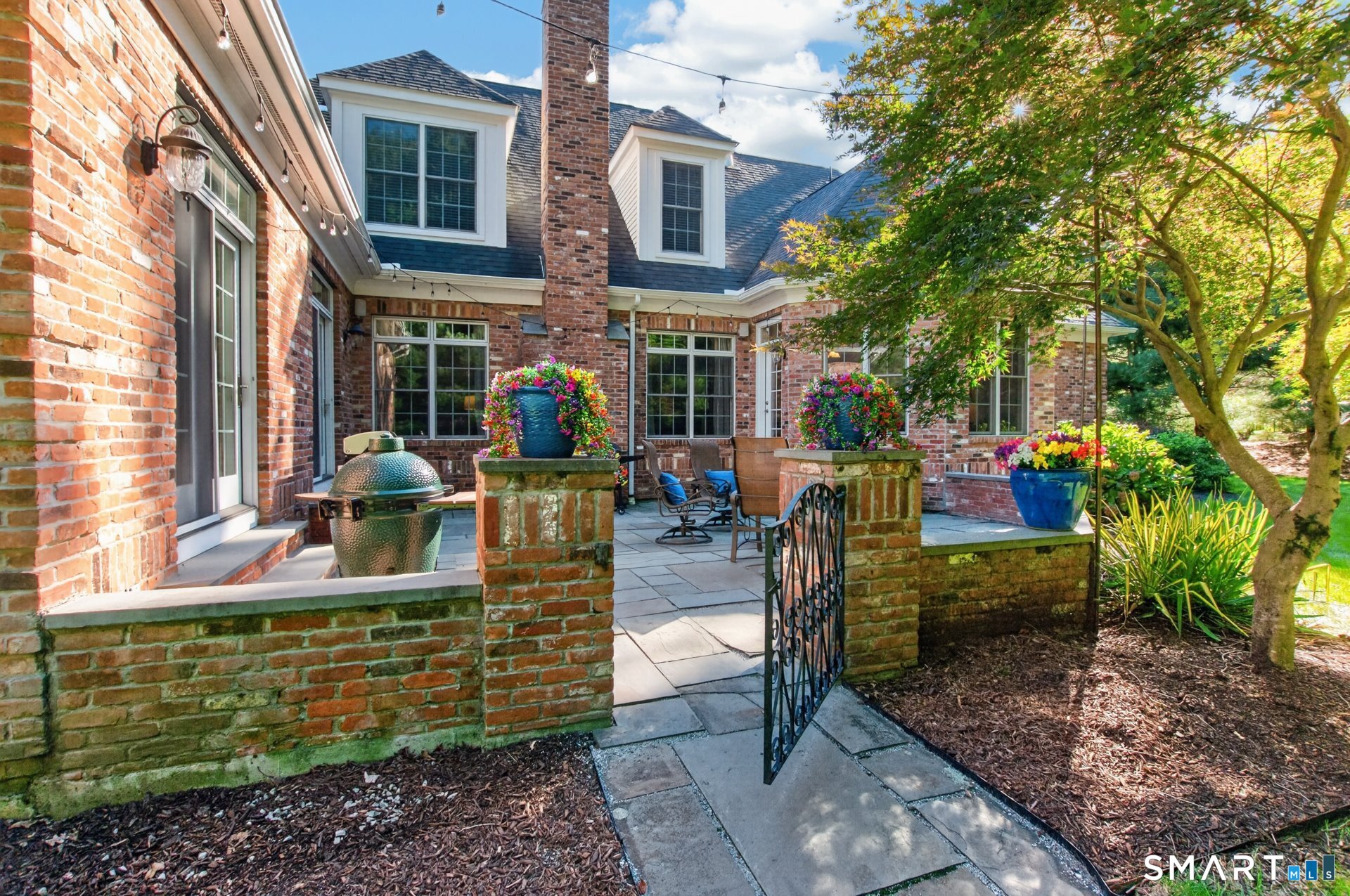 Brick home patio with stone pavers, outdoor seating, a ceramic grill, and colorful flower planters.