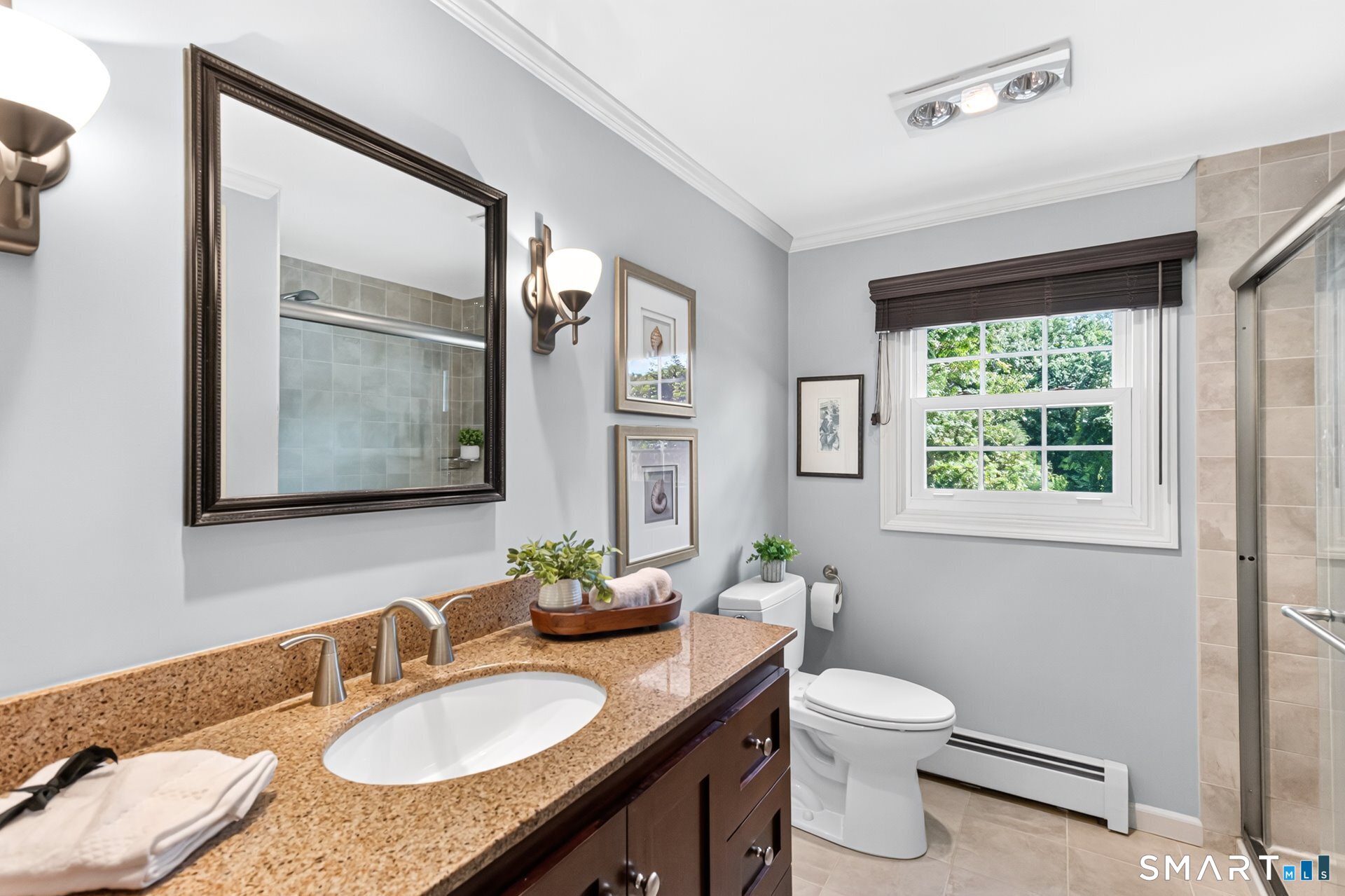 Bathroom with a granite vanity, dark wood cabinets, tiled glass shower, and window overlooking greenery.