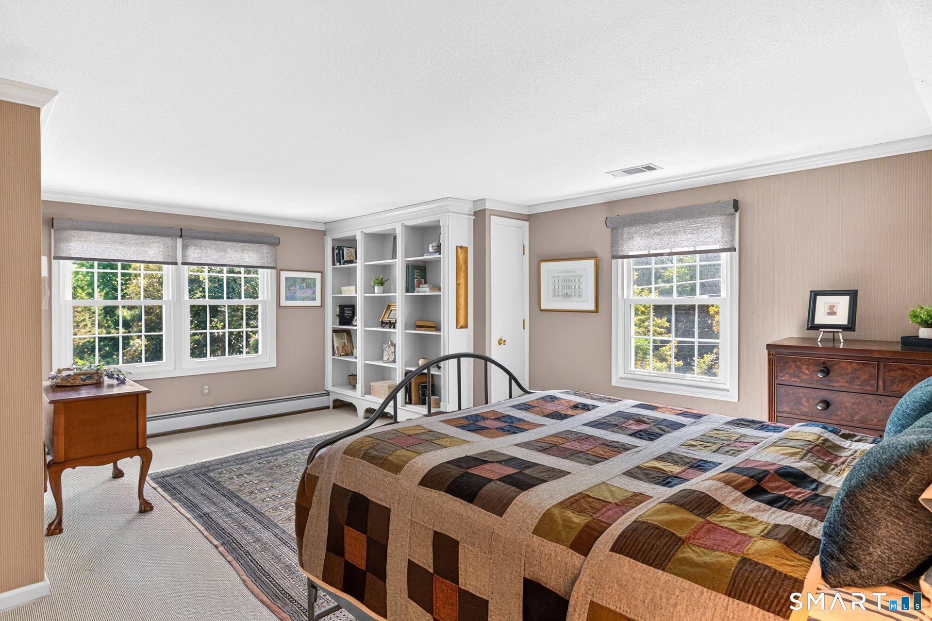 Furnished bedroom with a patchwork quilt, built-in bookshelves, and three windows overlooking greenery.