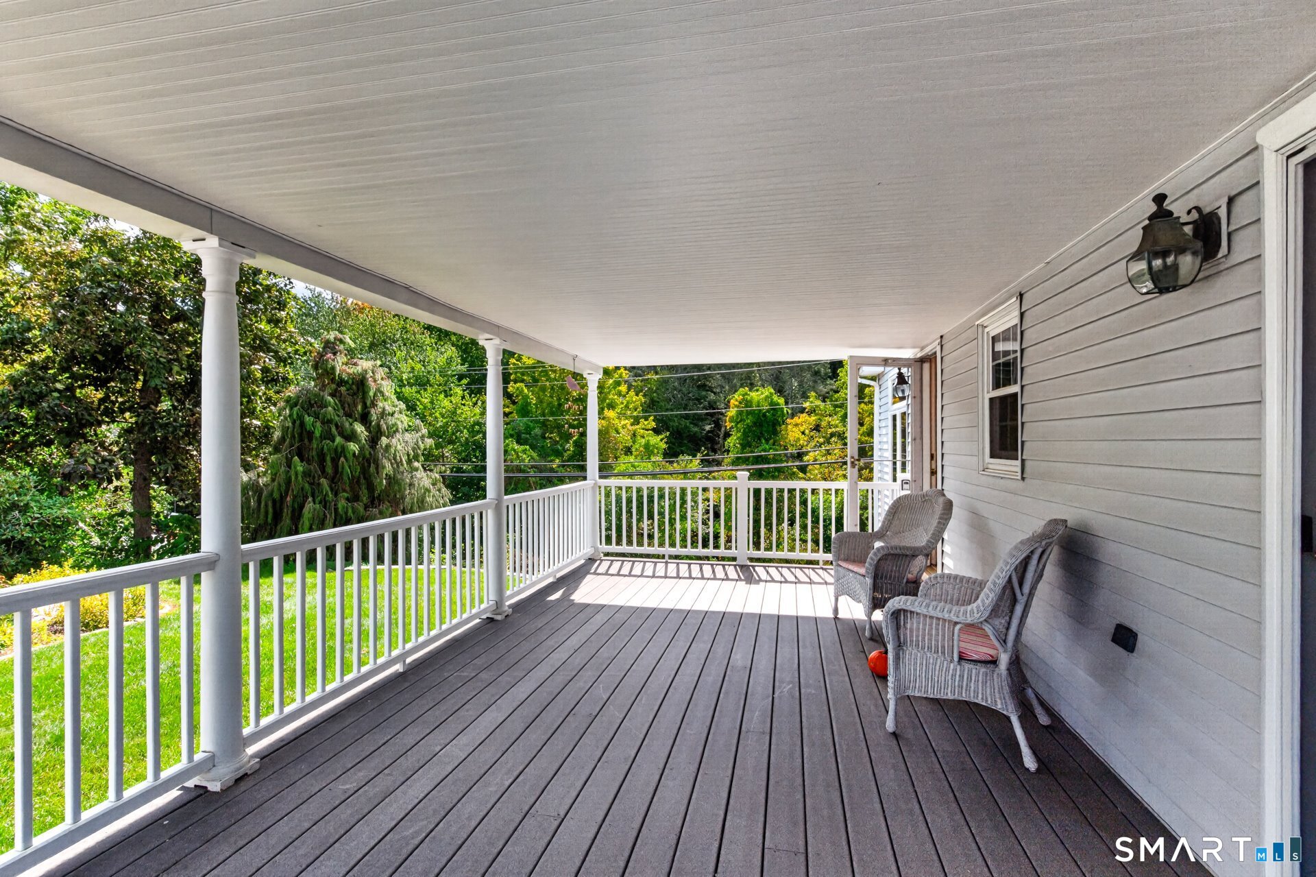 Covered porch with white railings, dark plank flooring, and wicker chairs overlooking leafy greenery.