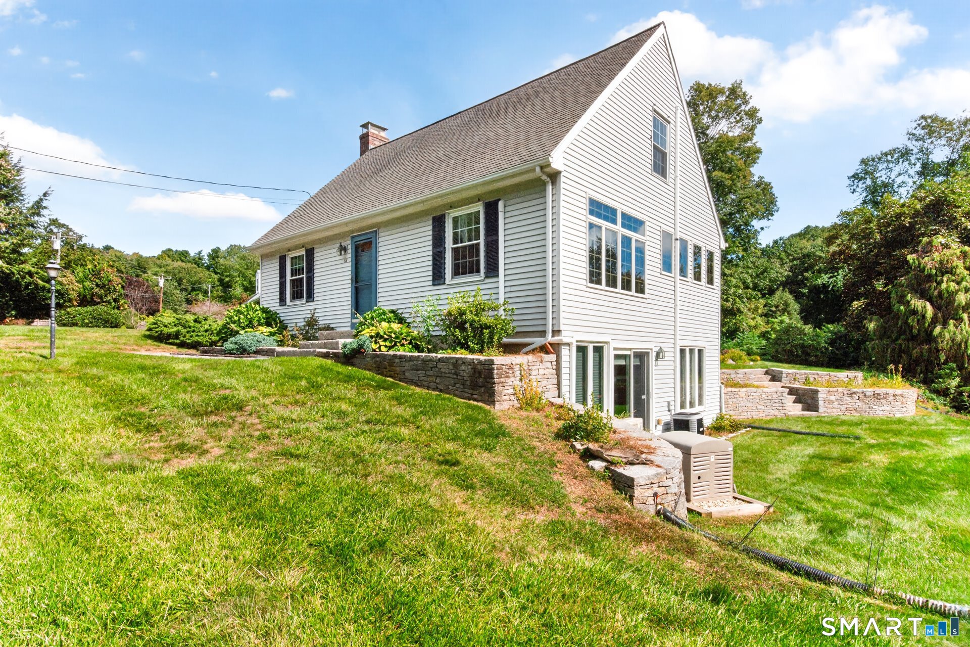 Light-sided house with a steep gabled roof, blue door, walkout lower level, and stone retaining walls.