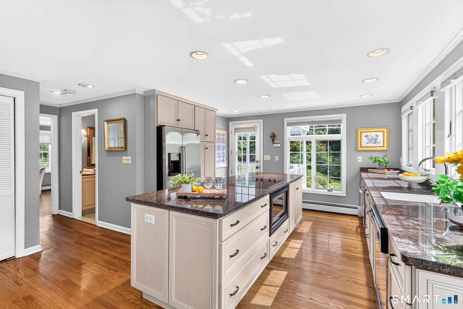 Kitchen with a large granite island, pale cabinets, hardwood floors, and windows overlooking greenery.
