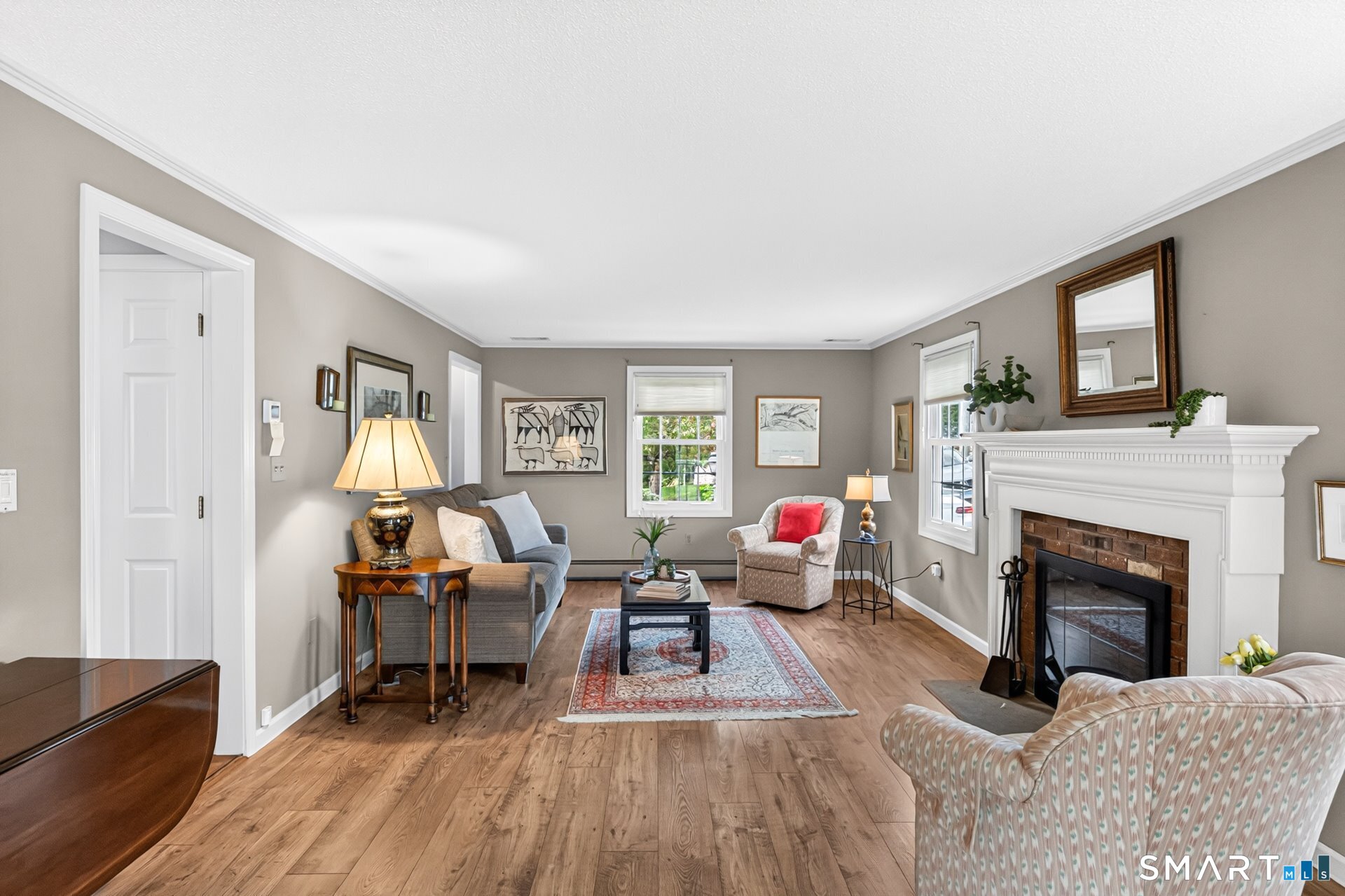 Staged living room with hardwood floors, a white fireplace, and seating around a patterned rug.