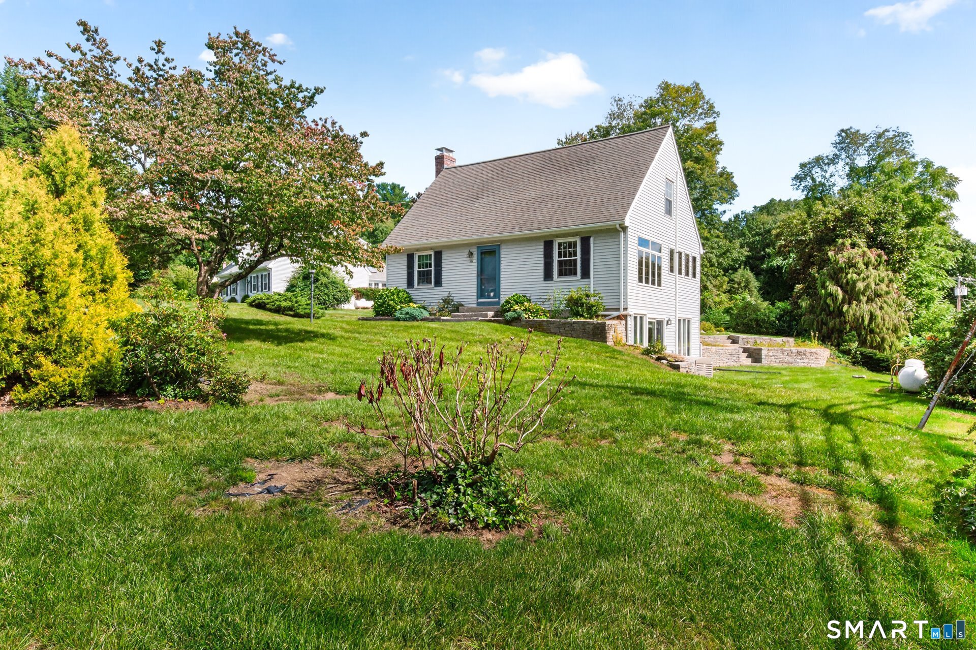 White-sided home with a steep gabled roof, teal front door, landscaped lawn, and stone retaining walls.