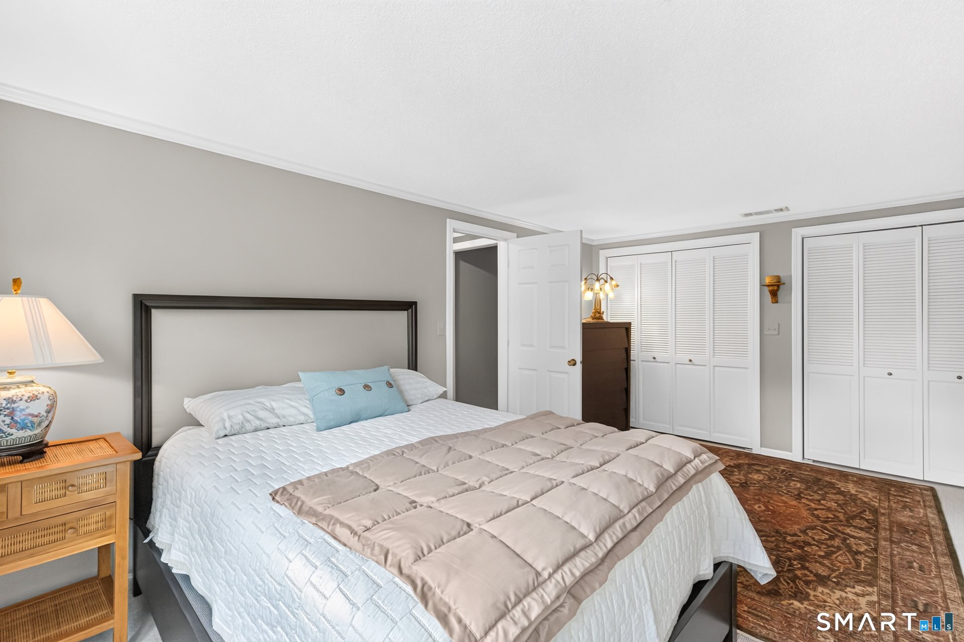 Furnished bedroom with a large white bed, beige quilt, louvered closets, and patterned rug.