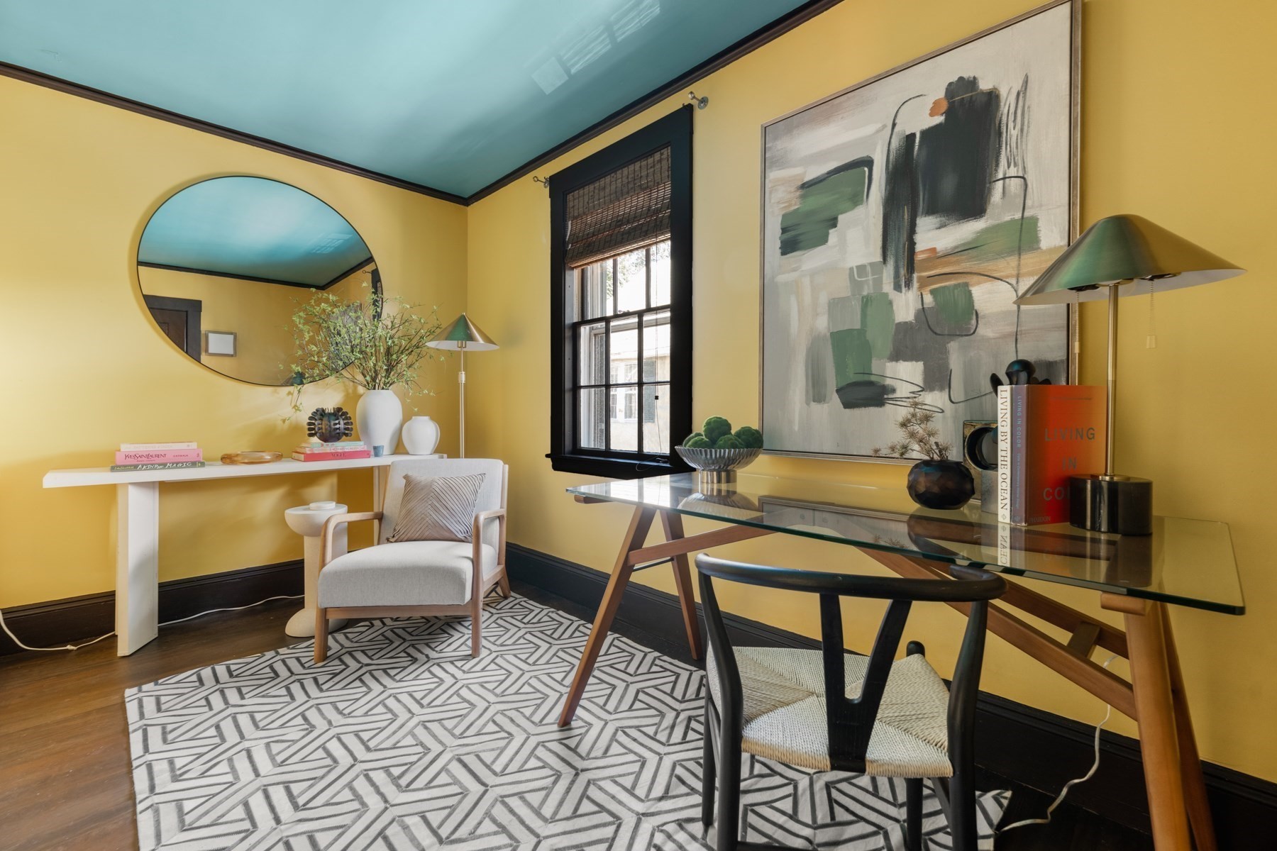 Furnished home office with mustard-yellow walls, teal ceiling, glass desk, and geometric rug.
