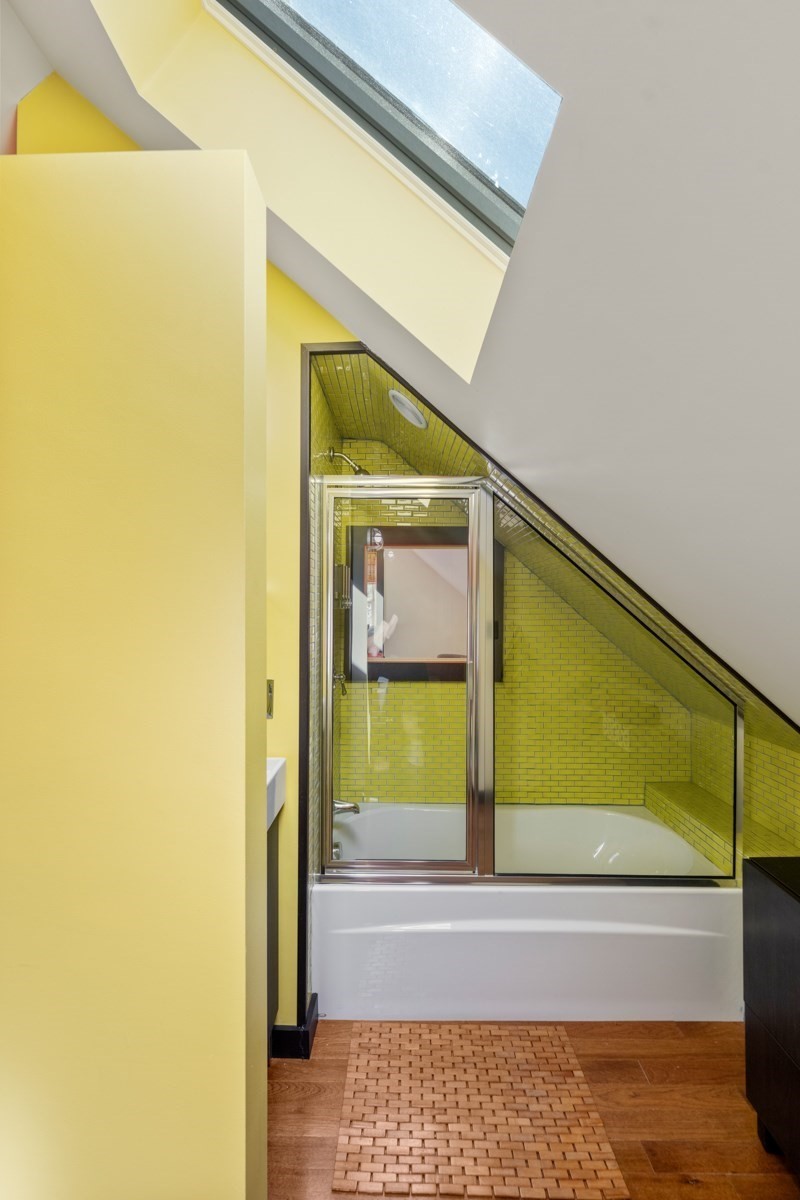 Bathroom with yellow-green tiled shower, white bathtub, sloped ceiling, and skylight.