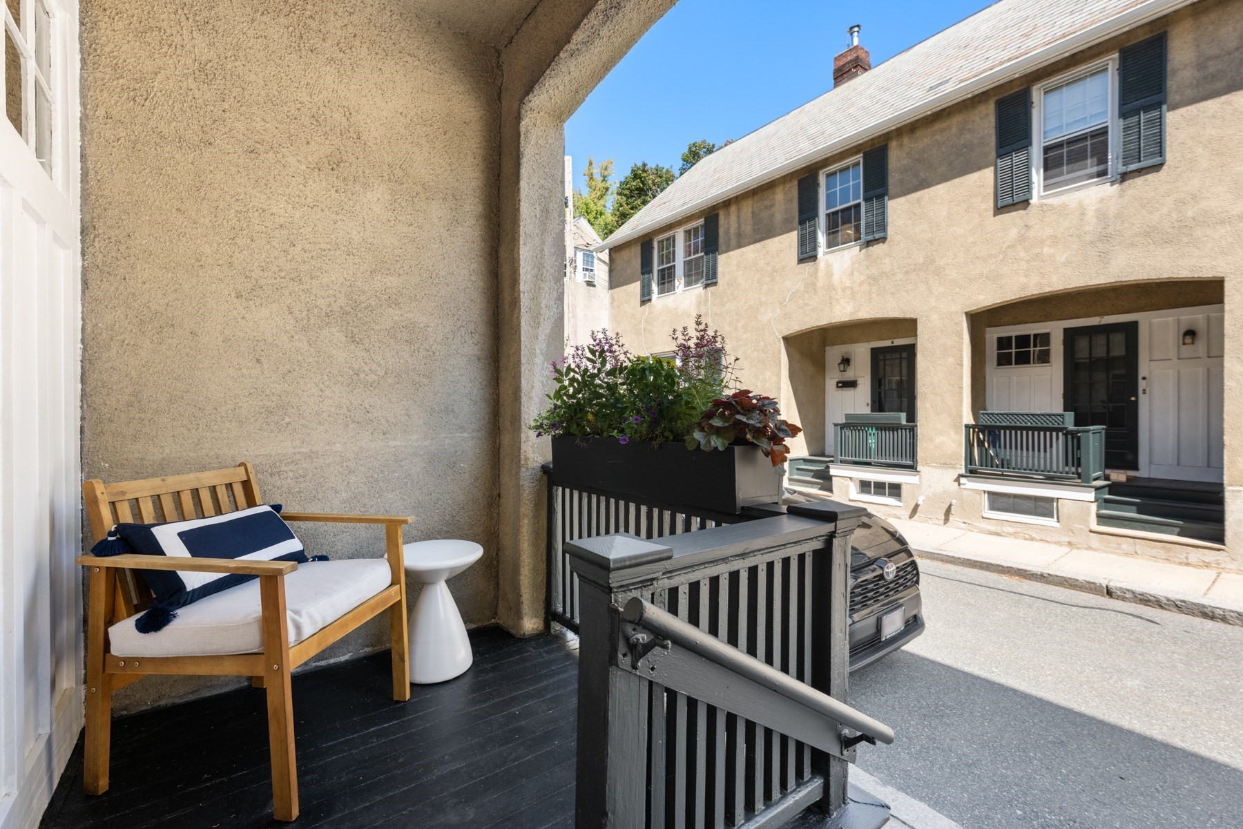 Furnished front porch with a cushioned wooden bench and flower planter facing a beige townhouse.