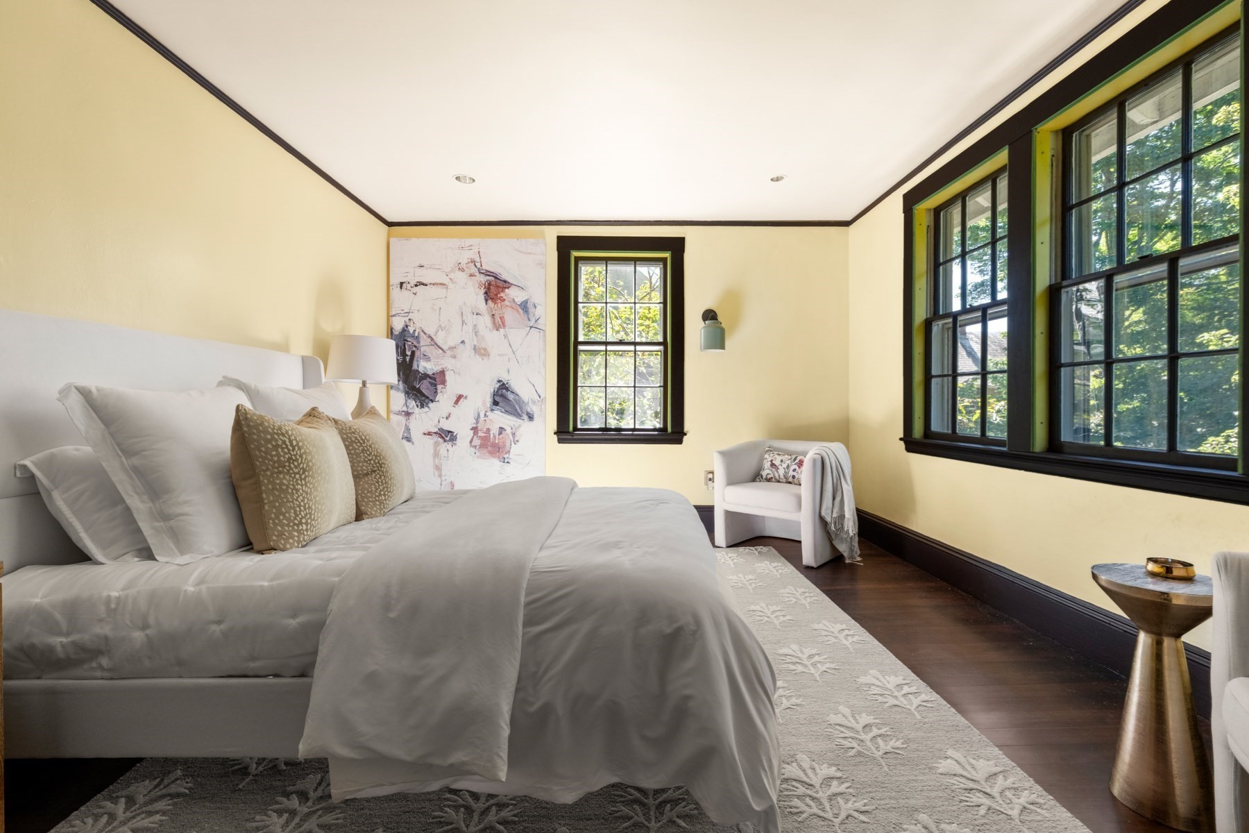 Bedroom with a white bed, abstract artwork, and tall black-framed windows overlooking leafy trees.