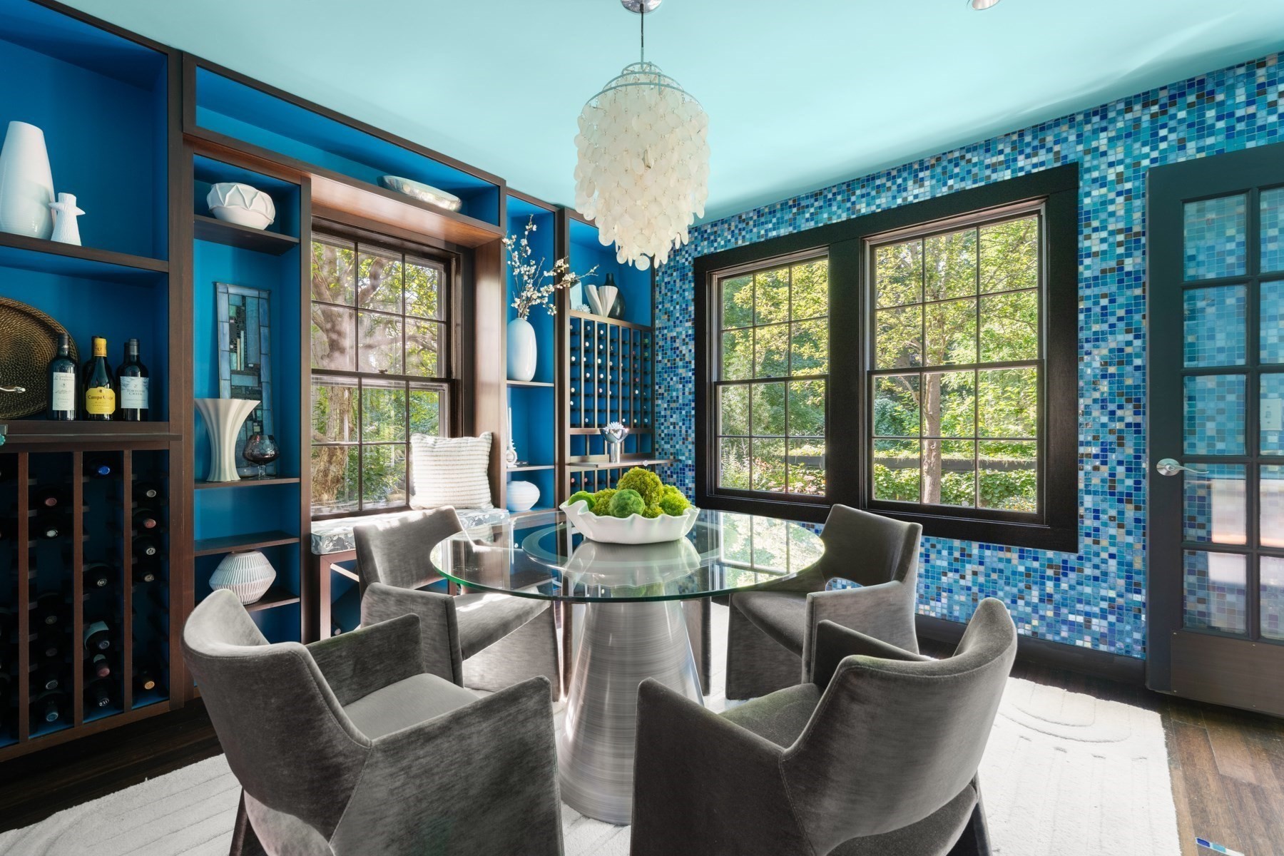 Dining room with blue mosaic tile walls, built-in wine racks, and a round glass table by large windows.