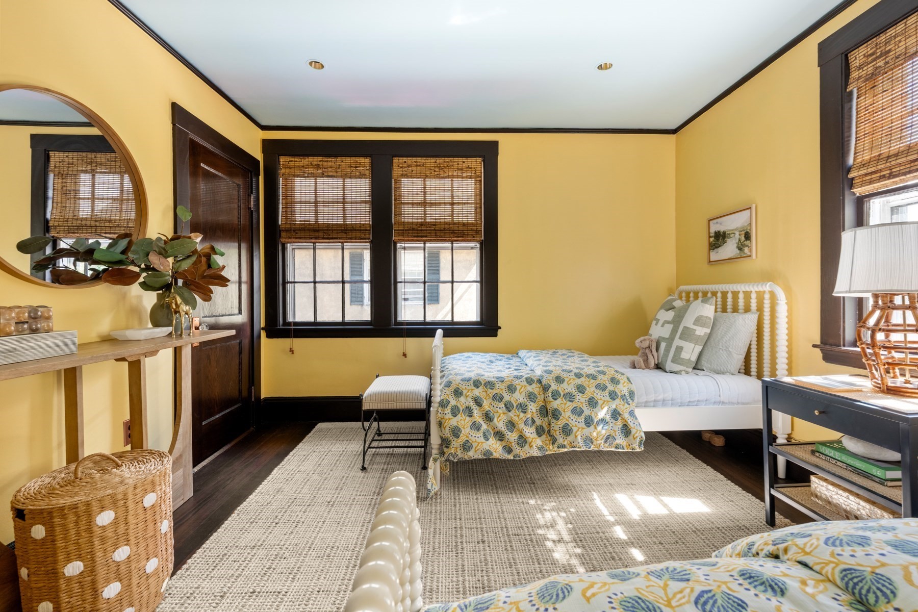 Bedroom with mustard-yellow walls, dark trim, bamboo shades, and white beds with patterned bedding.