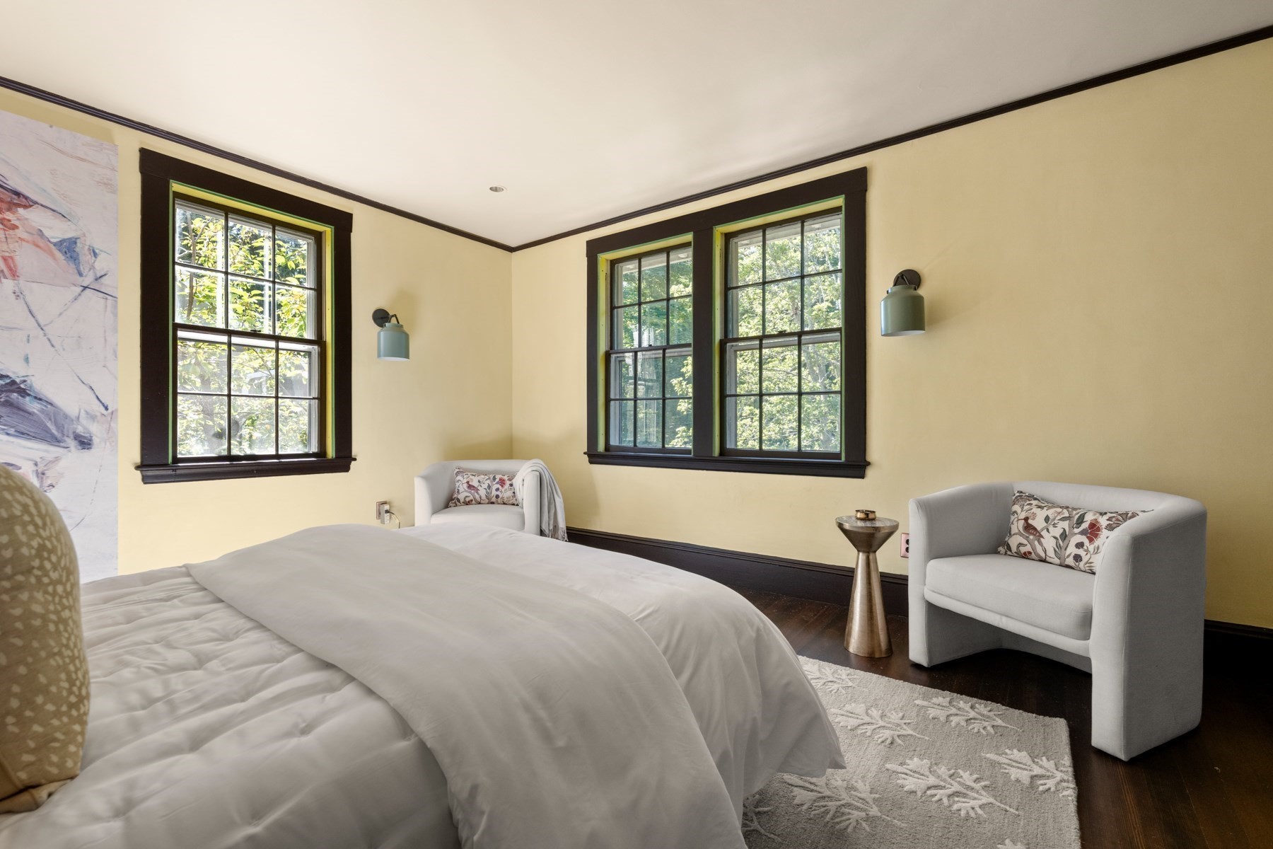 Staged bedroom with white bedding, dark-trimmed windows, and gray upholstered chairs.