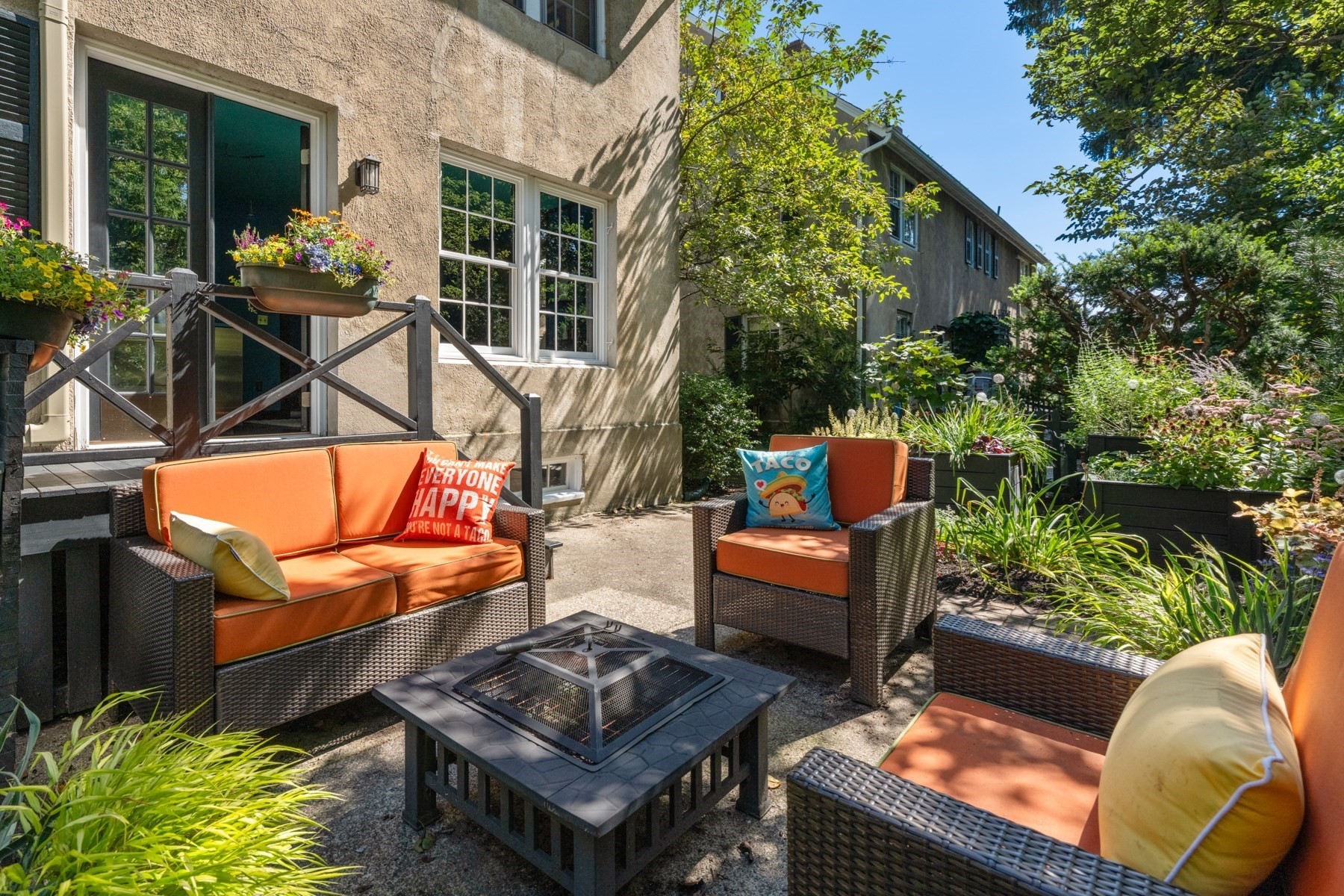 Garden patio with orange-cushioned wicker seating, a square fire pit, and raised flower beds.