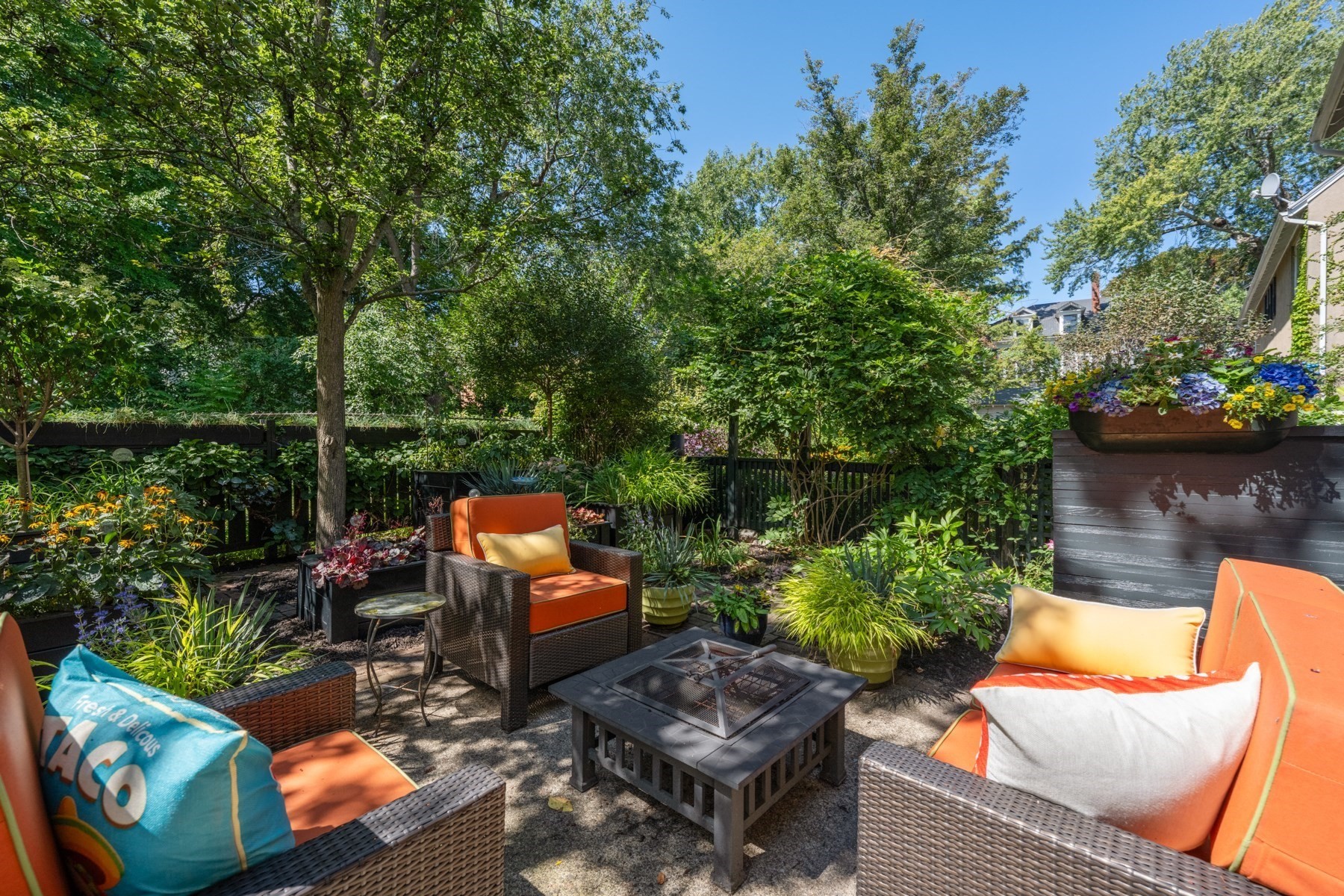 Garden patio with orange-cushioned wicker seating, a fire pit table, and lush landscaping.