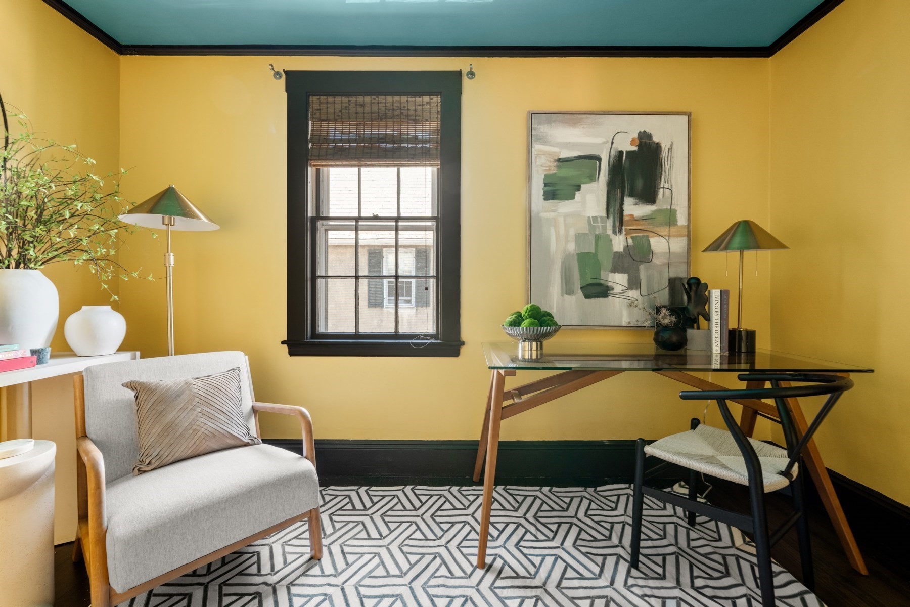 Home office with mustard-yellow walls, dark trim, glass desk, abstract art, and geometric rug.