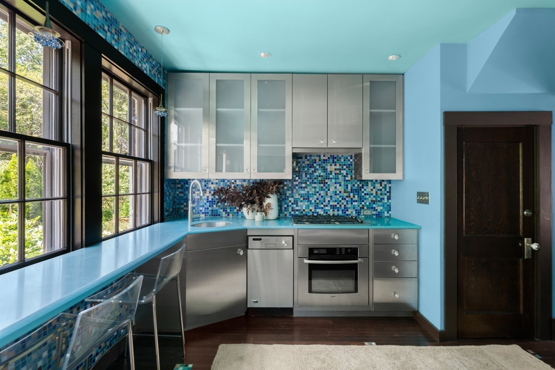 Modern kitchen with turquoise counters, blue mosaic backsplash, stainless appliances, and large windows.