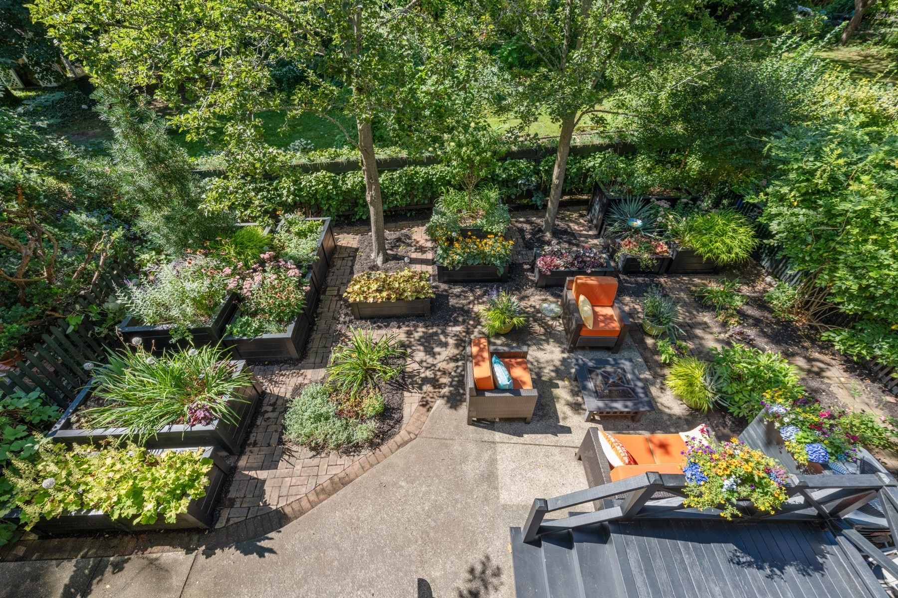 Garden patio with raised flower beds, brick paths, and orange-cushioned seating beneath leafy trees.