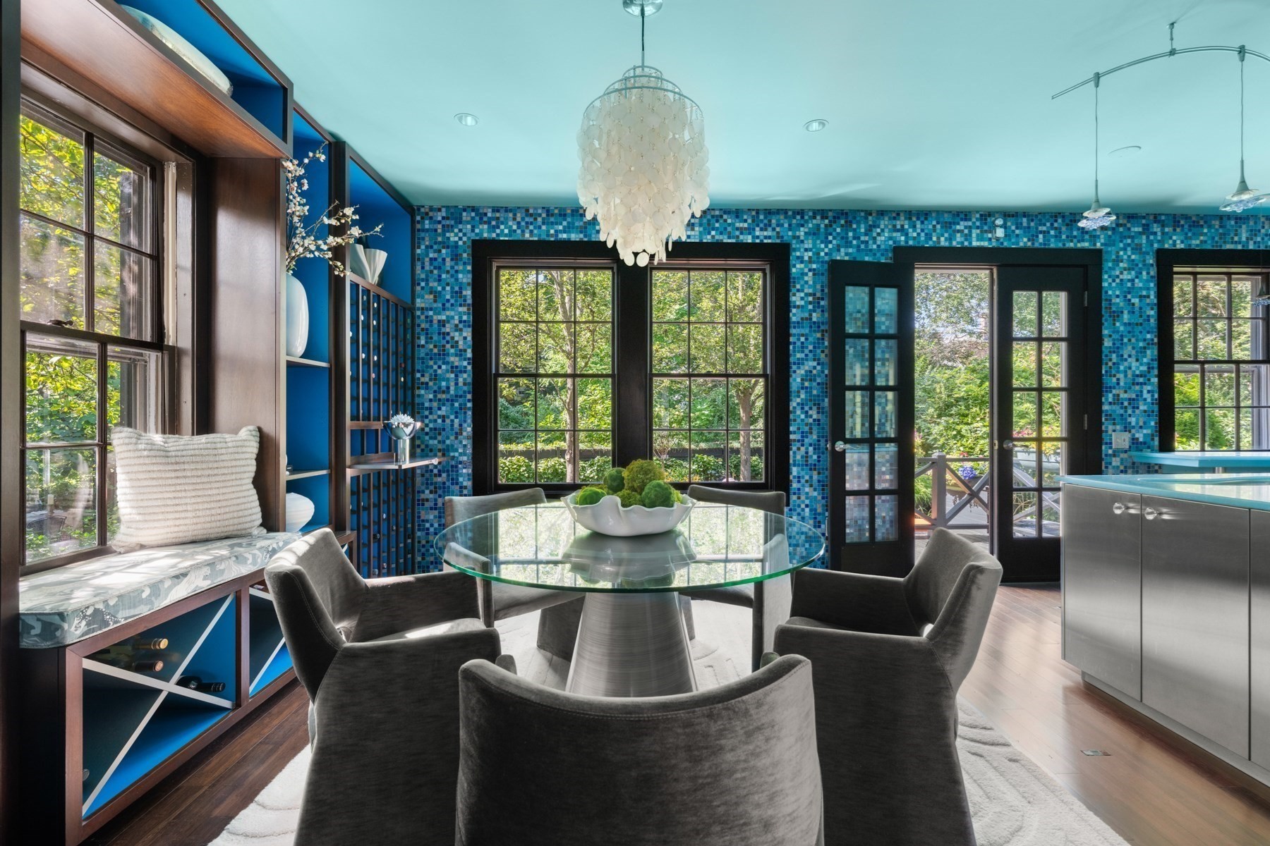 Dining room with blue mosaic tile walls, a glass table, sculptural chandelier, and garden-facing windows.