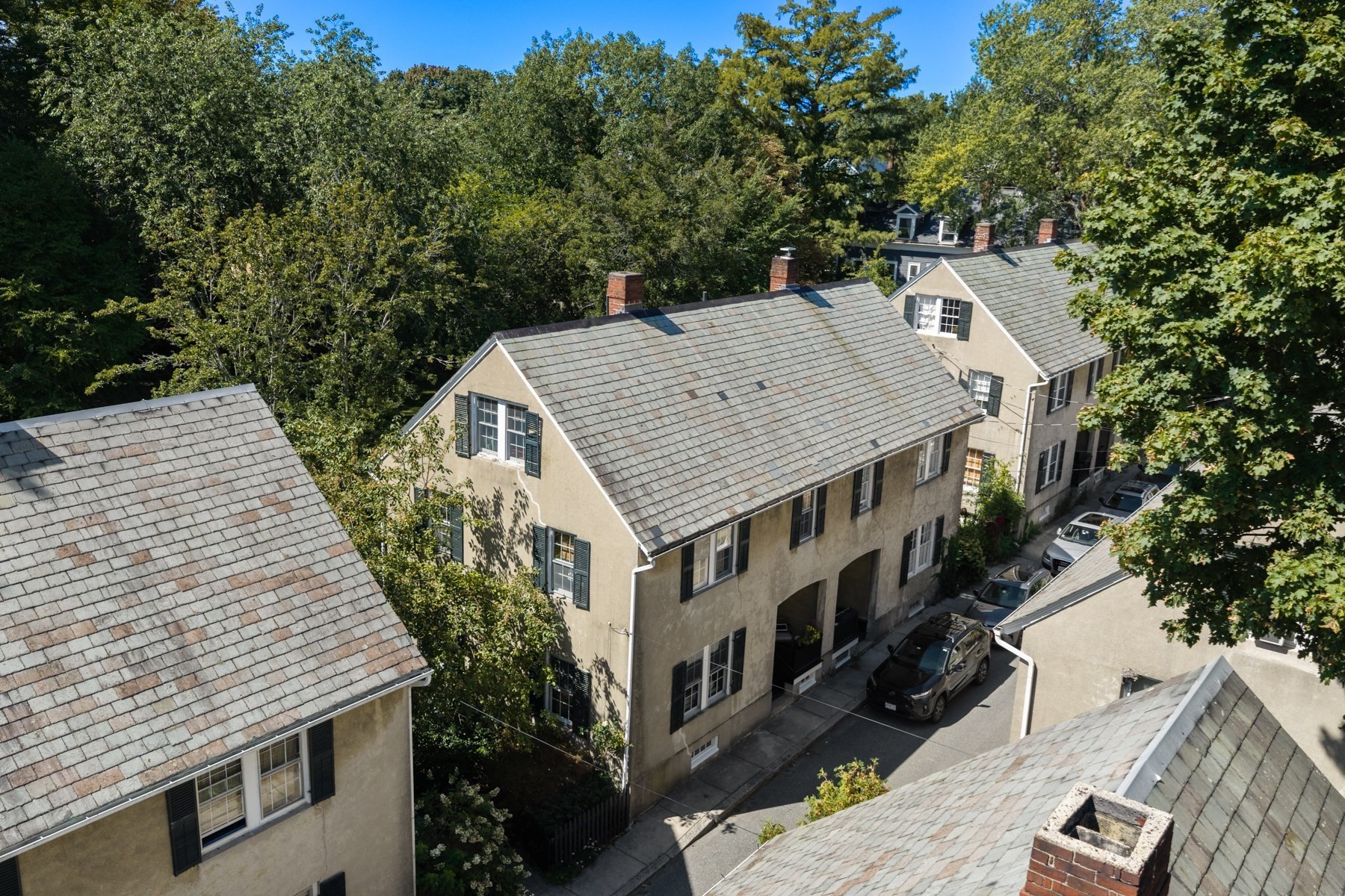 Aerial view of a beige multi-unit building with a long shingle roof, brick chimneys, and a narrow parking lane.