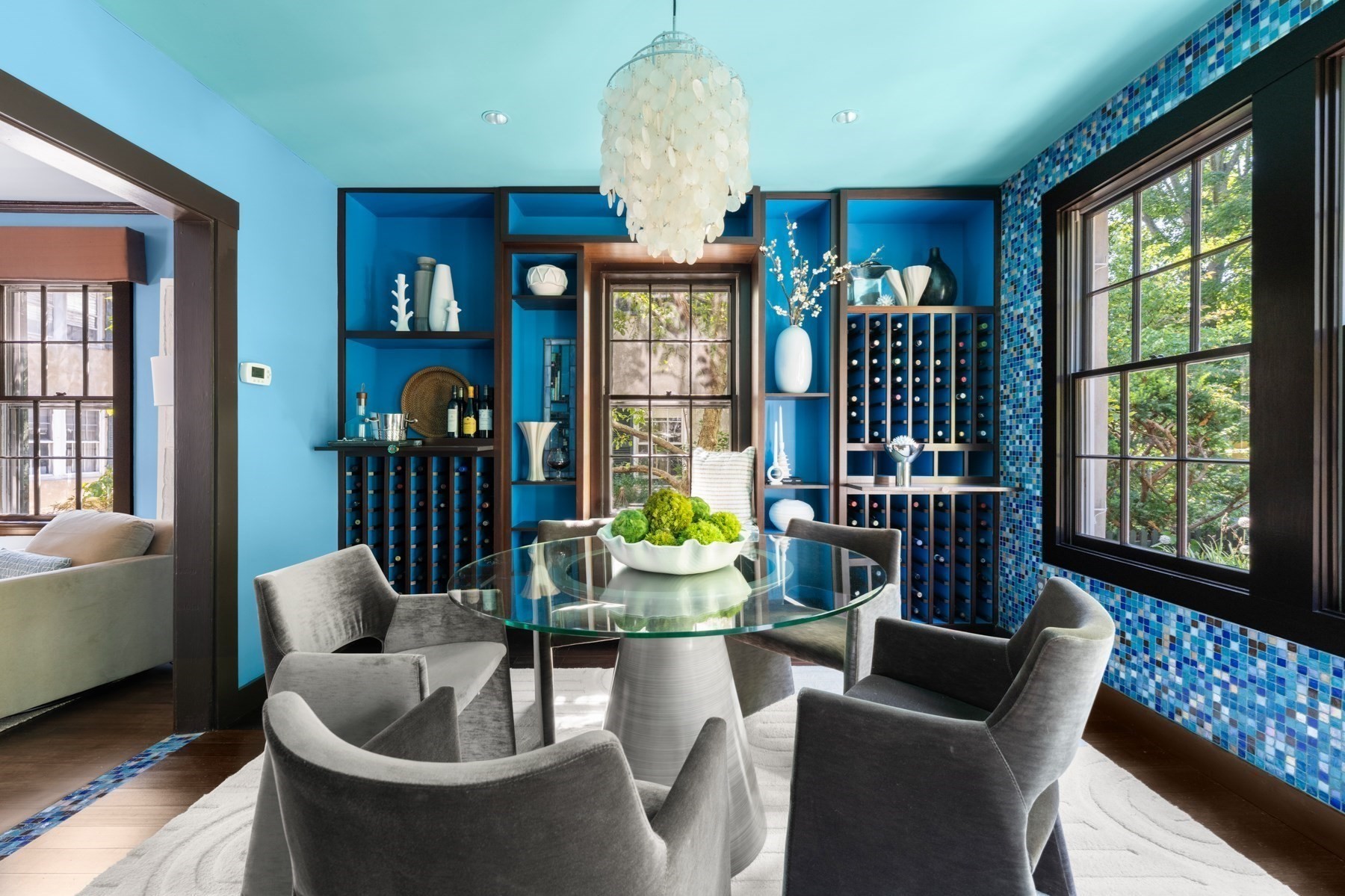 Dining room with blue wine-storage built-ins, mosaic tile, and a glass table beneath a shell chandelier.