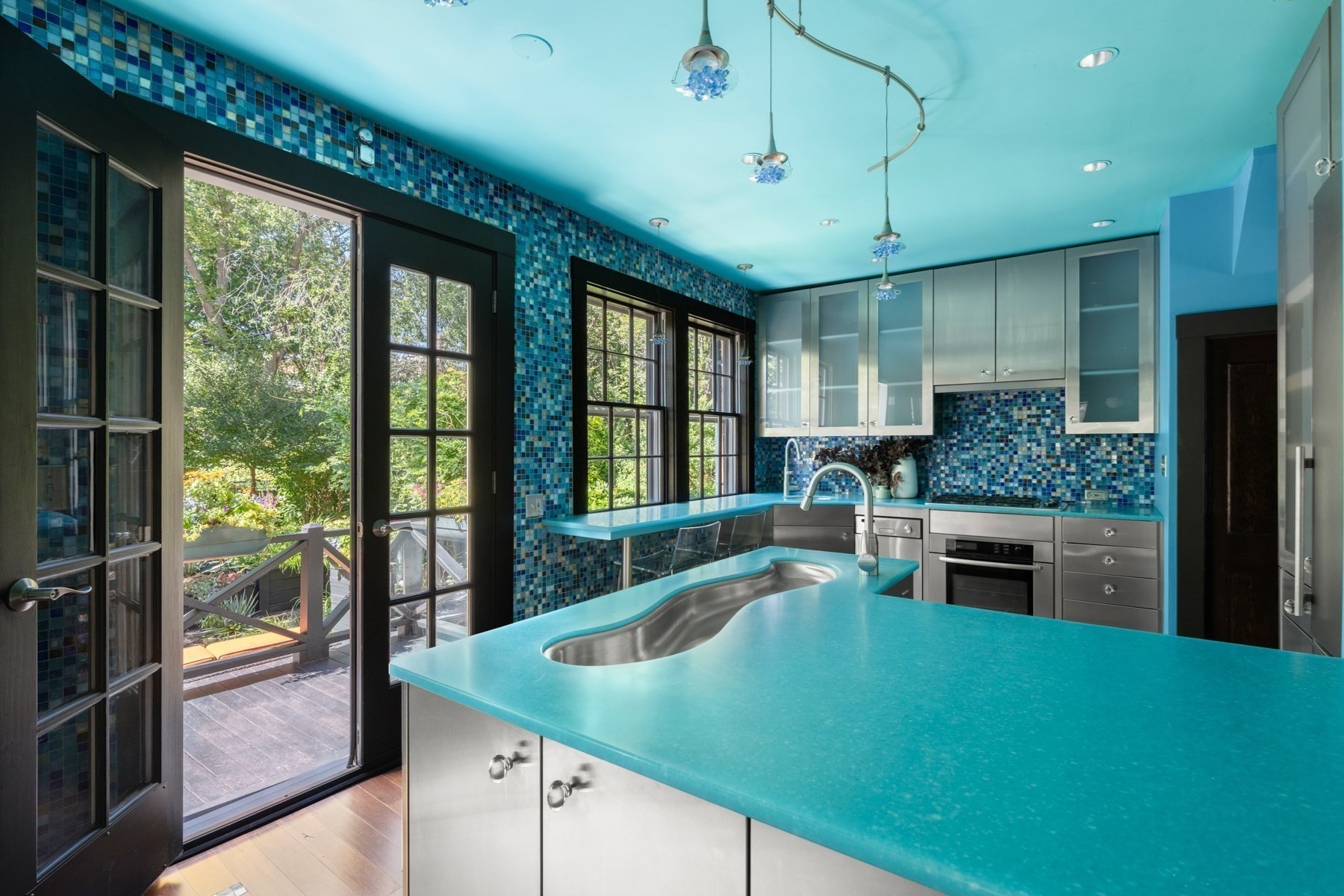 Modern kitchen with turquoise counters, blue mosaic tile, stainless steel cabinetry, and glass doors to a garden deck.