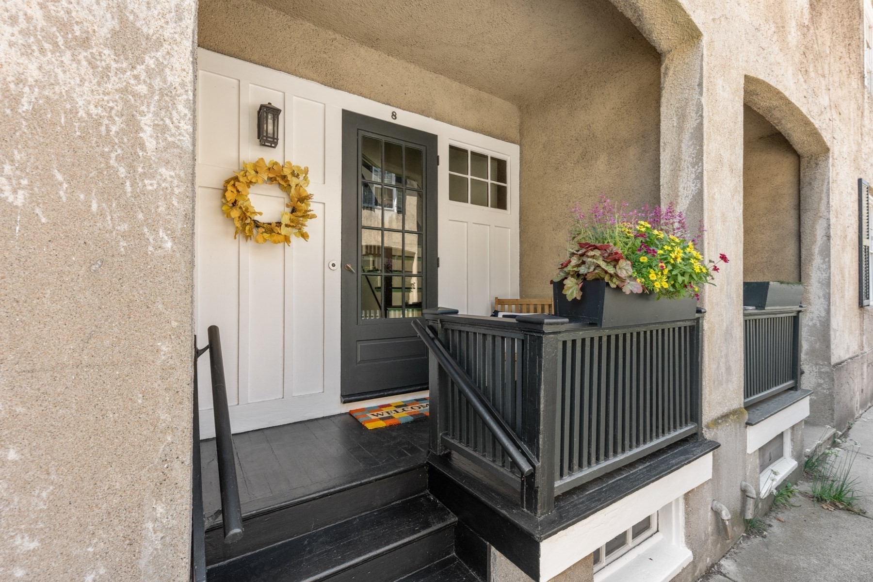 Home entrance with a black glass-paneled door, yellow wreath, and flower-filled planter on the porch railing.