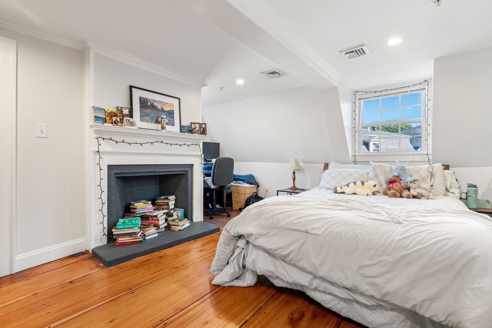 Bedroom with a white fireplace, books, sloped ceiling, and warm hardwood floors.