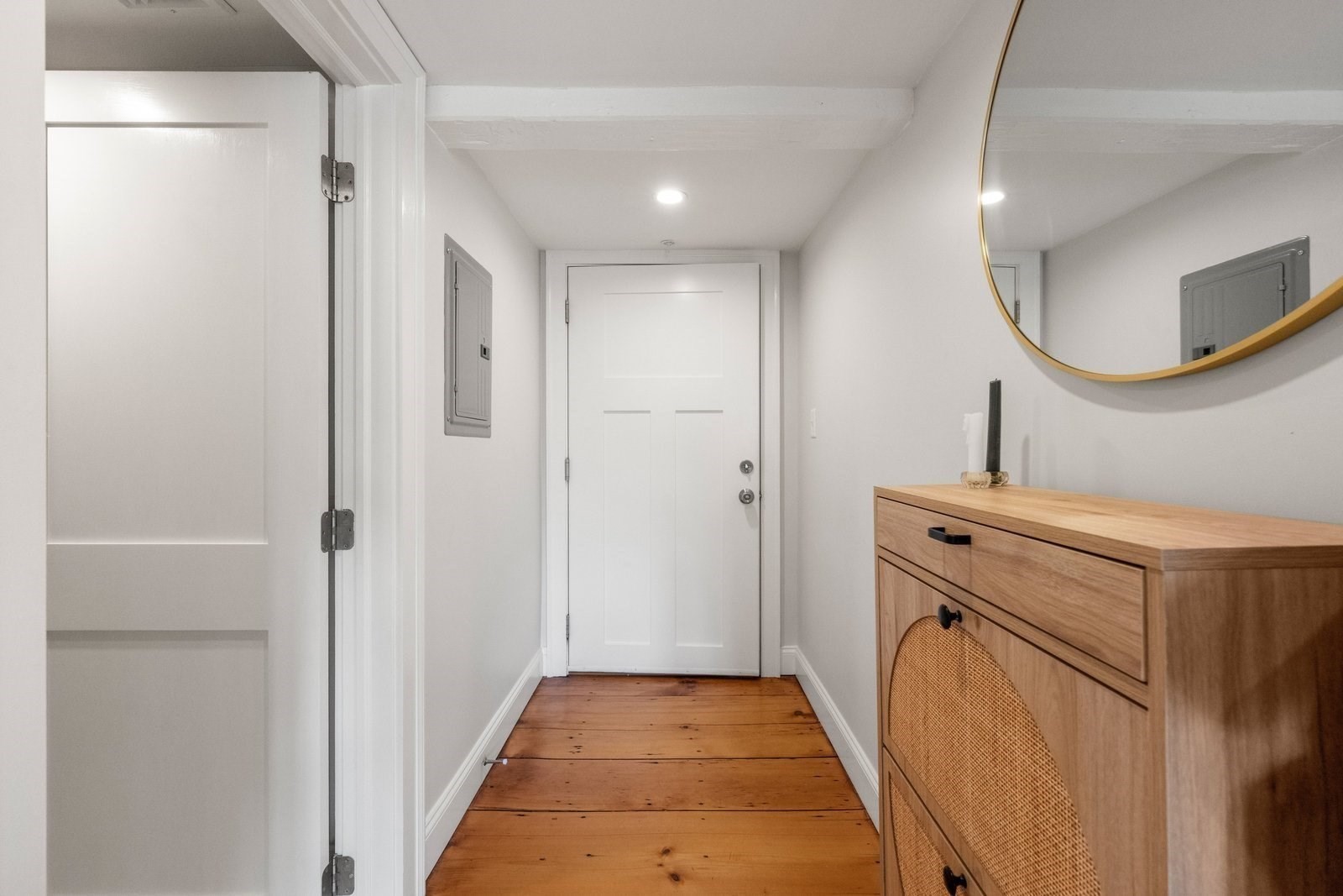 Entry hallway with a white door, warm plank flooring, cane-front cabinet, and round mirror.