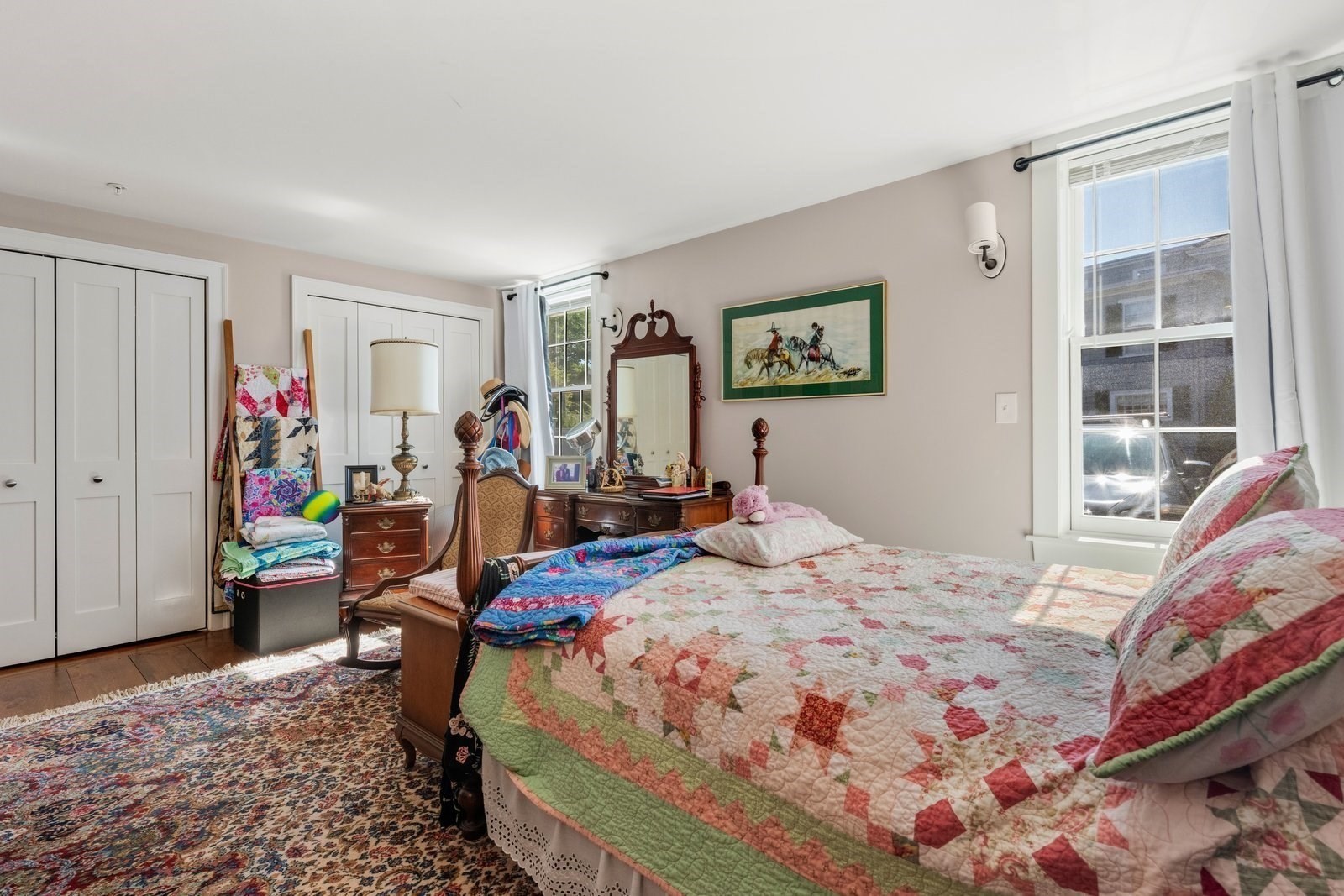 Furnished bedroom with a patchwork quilt, antique dresser and mirror, and bright window.