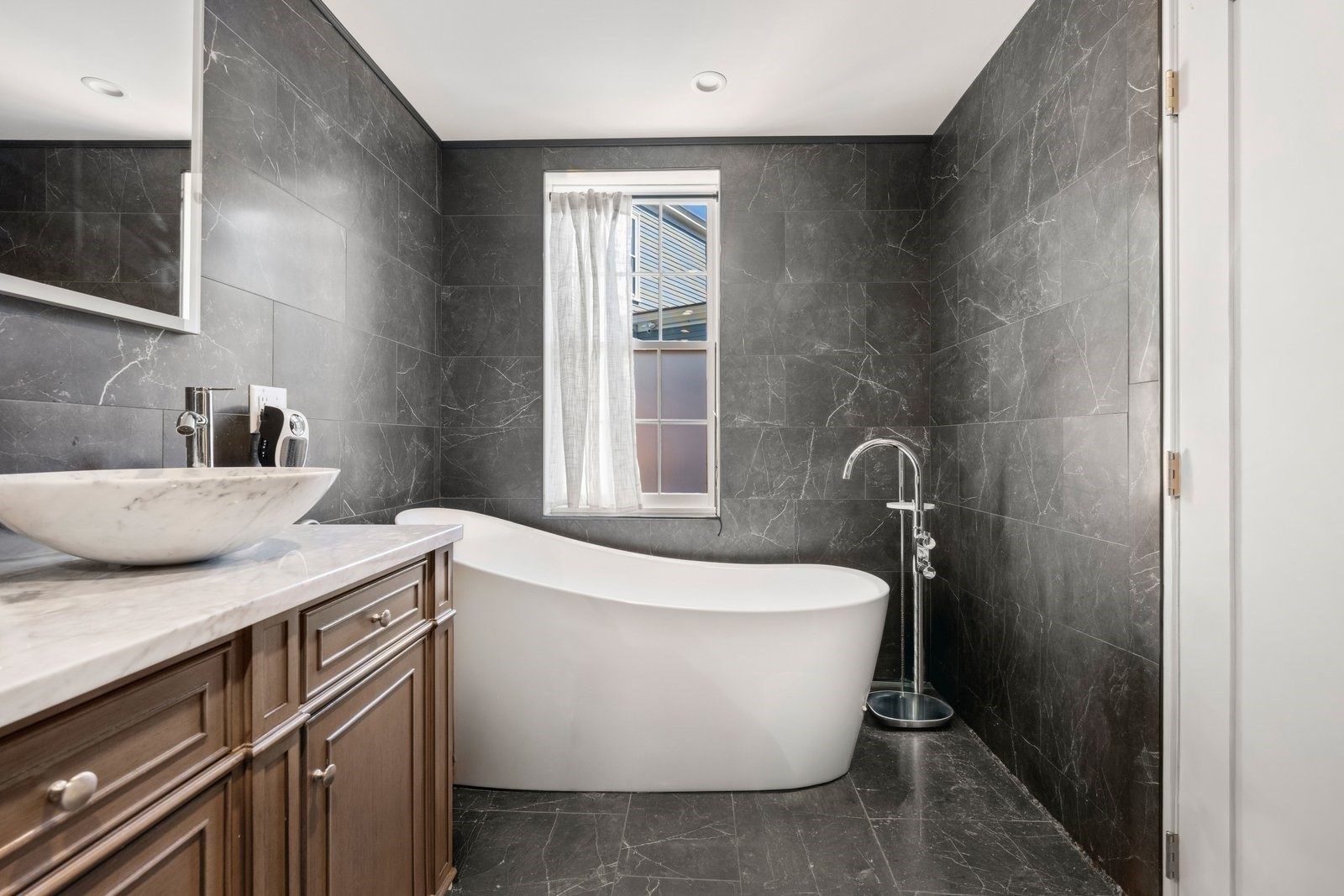 Bathroom with a freestanding soaking tub, dark marble-pattern tile, and a marble-topped wood vanity.