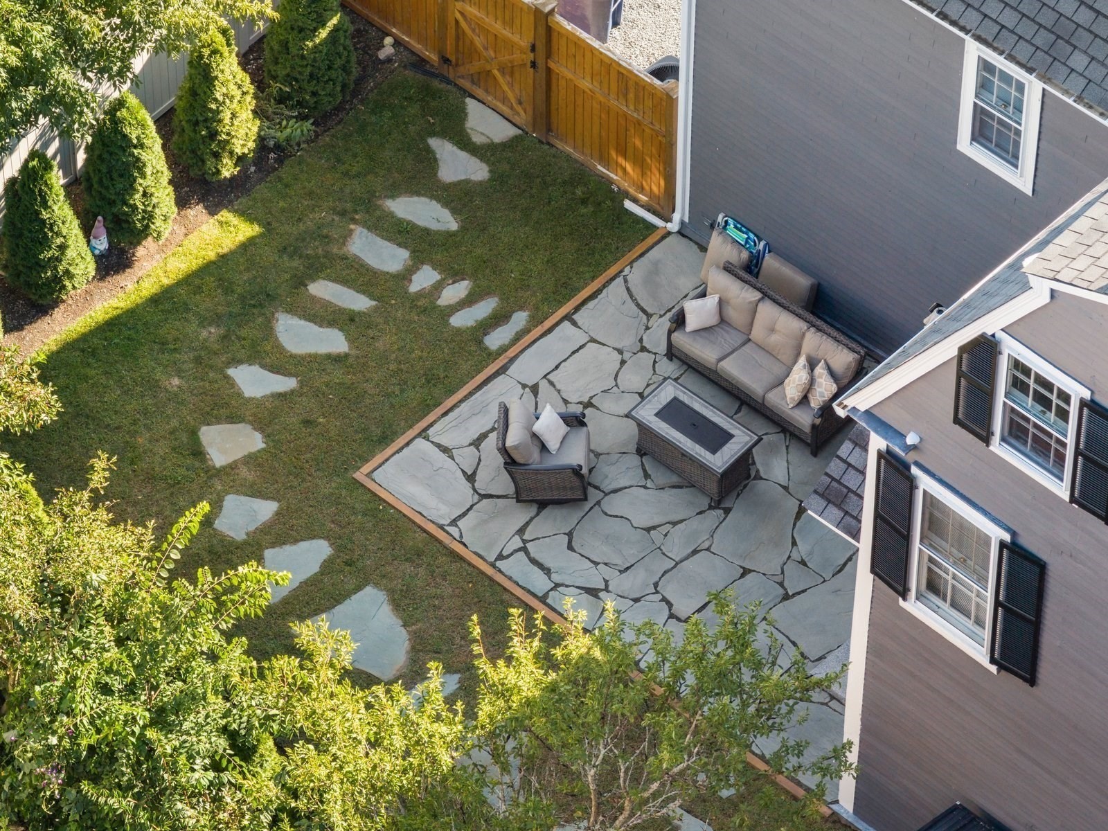 Aerial backyard view with a furnished flagstone patio, fire table, stepping-stone lawn, and privacy fence.