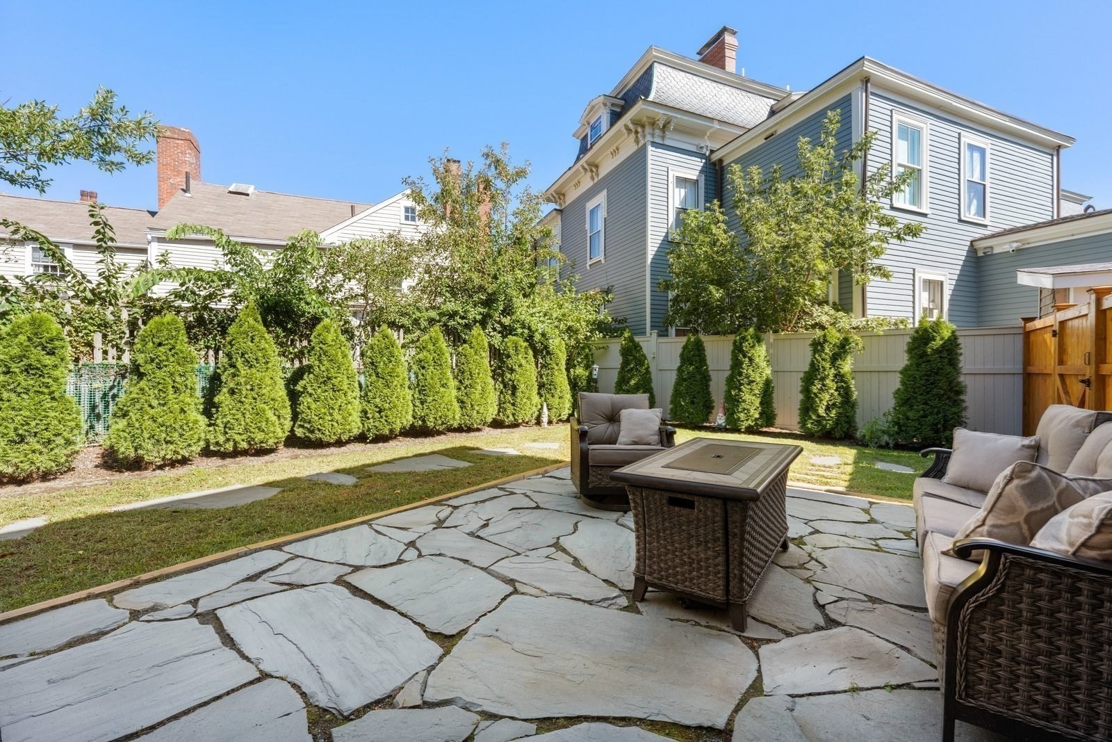 Slate patio with wicker seating in a fenced backyard beside a blue-gray house.
