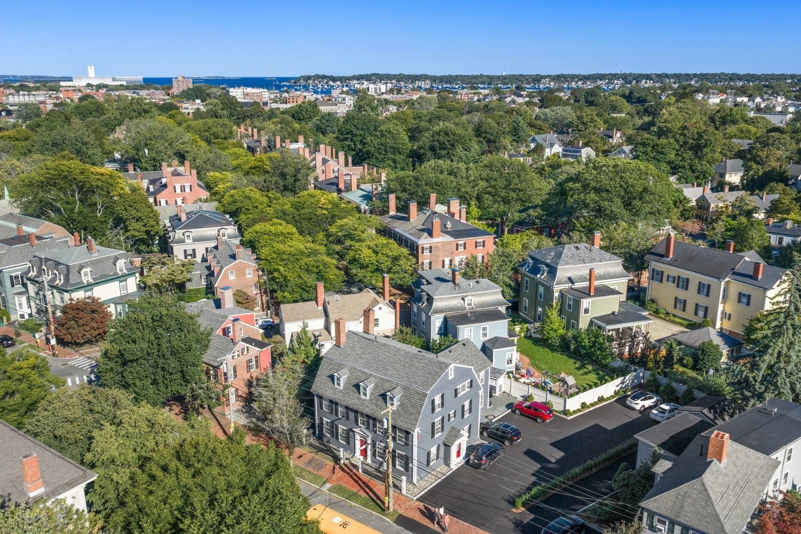 Aerial view of leafy homes and neighborhood streets, with a harbor in the distance.